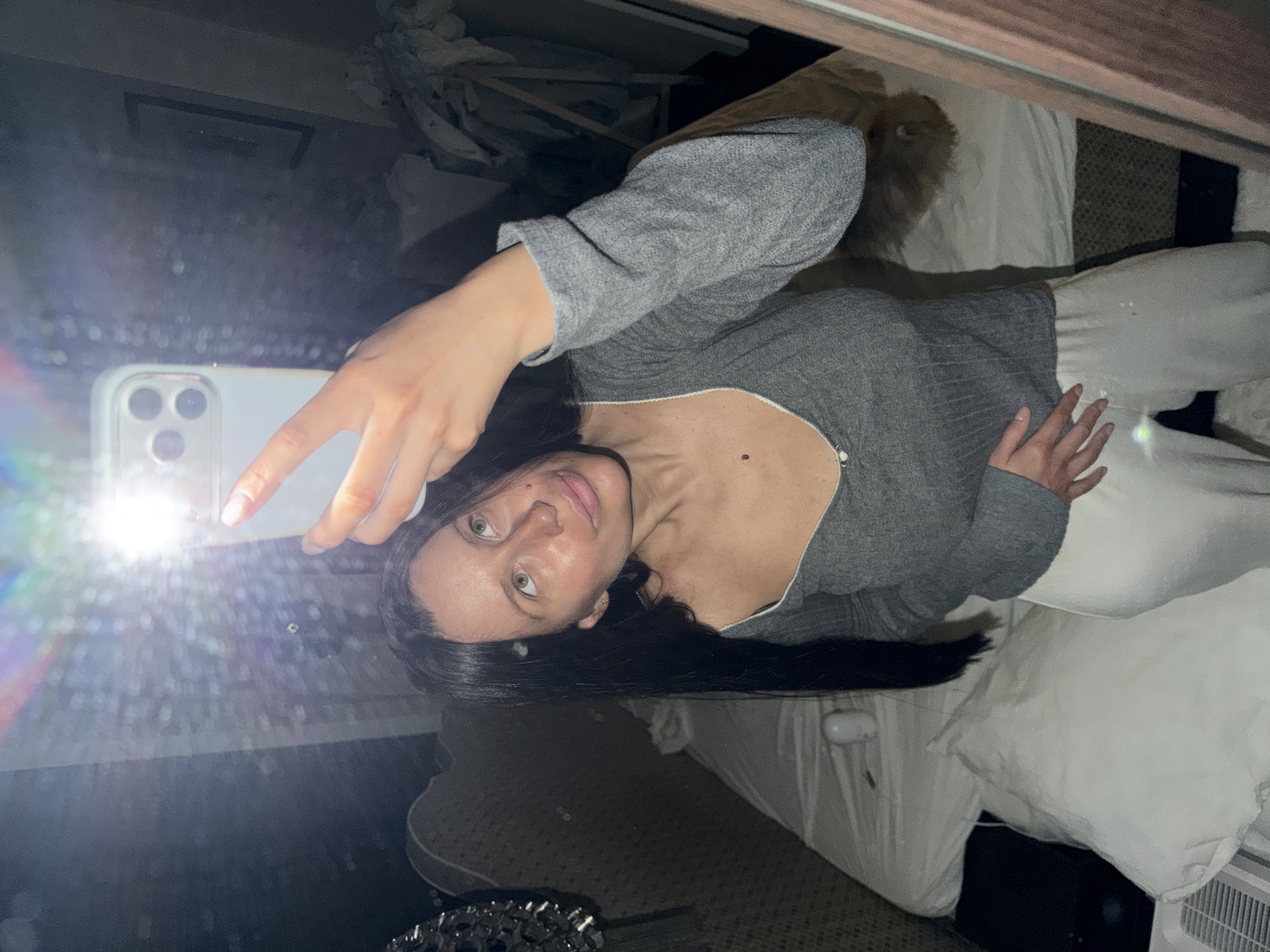 A woman is taking a selfie in a mirror. She is holding a white smartphone in her left hand. She is wearing a gray long-sleeved shirt and white pants. Her hair is black. The background appears to be a bedroom with a bed and possibly a dresser. There's no evidence to suggest the image was taken in Khimki, Russia.