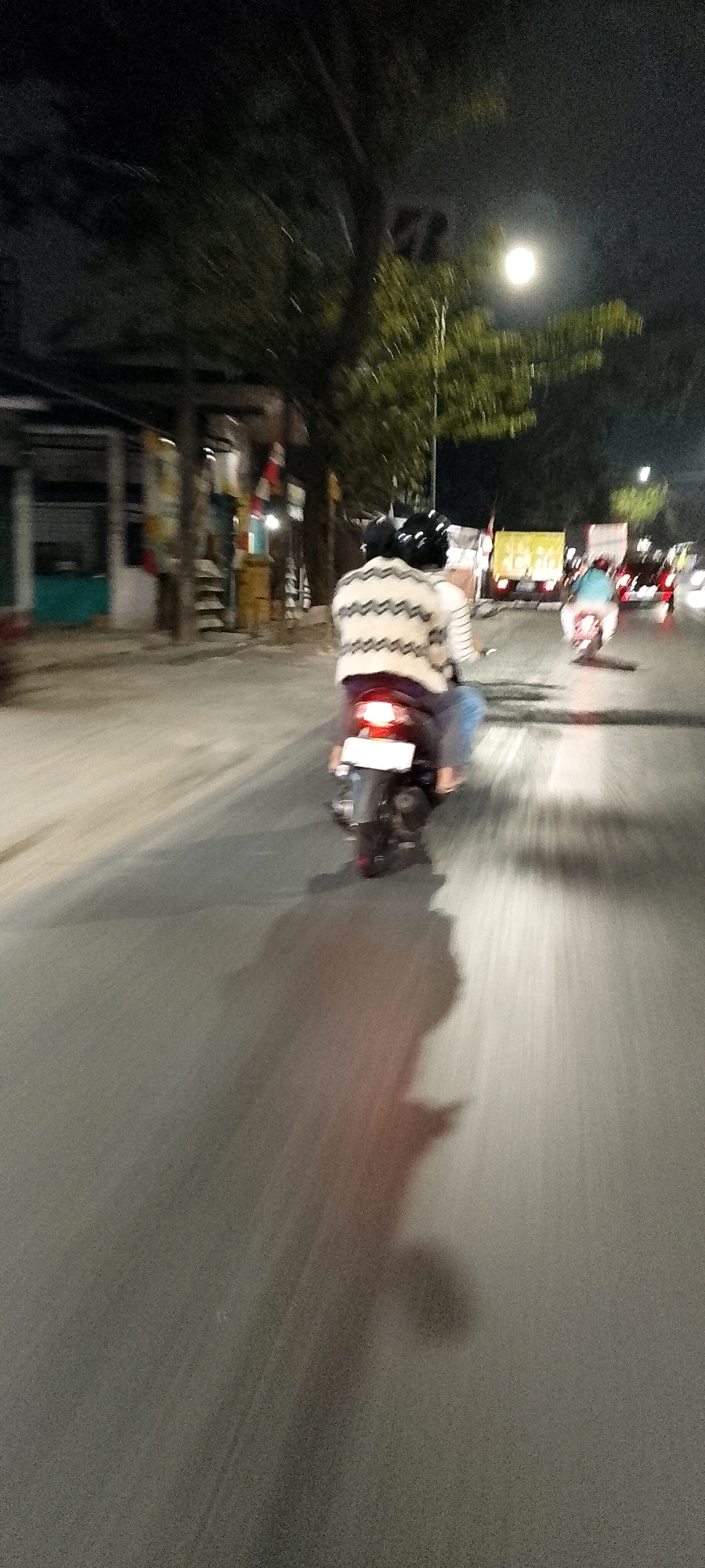 The image shows a slightly blurred nighttime scene on a street in Bekasi, Indonesia. In the foreground, two people are riding a scooter on the asphalt road. The person in the back is wearing a striped sweater and a black helmet. The scooter's taillight is illuminated, casting a reddish glow on the road. A shadow of the scooter and its riders is visible on the street. In the background, there are buildings and trees, and a bright light source, possibly a streetlamp or the moon, is visible through the foliage. Other vehicles, including a truck and possibly motorcycles, are also visible further down the road. The overall impression is a snapshot of everyday life on the move in an urban environment at night.