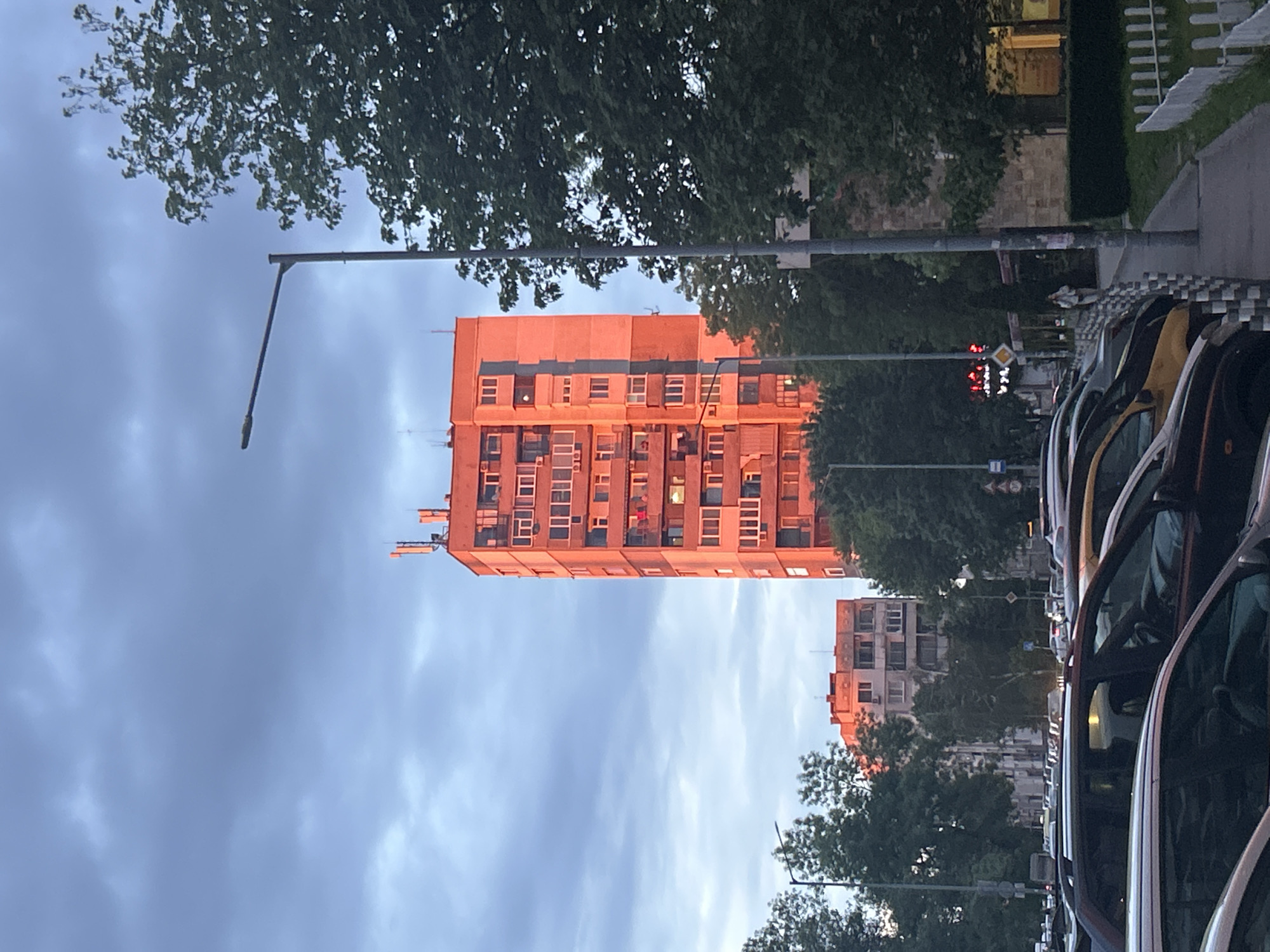 The image depicts a cityscape, likely in Sofia, Bulgaria, at dusk. A tall, rectangular apartment building dominates the center, illuminated by a warm, reddish-orange light, possibly from the setting sun. The building has multiple rows of windows and some antenna structures on its roof. Above and around the building are green trees and a cloudy sky. A street light is positioned to the left of the building. To the right, parked cars line the street, with their windshields reflecting the surrounding light. Further down the street, another similar building can be seen in the distance, also illuminated with the same warm light. A traffic light is visible, showing red lights.