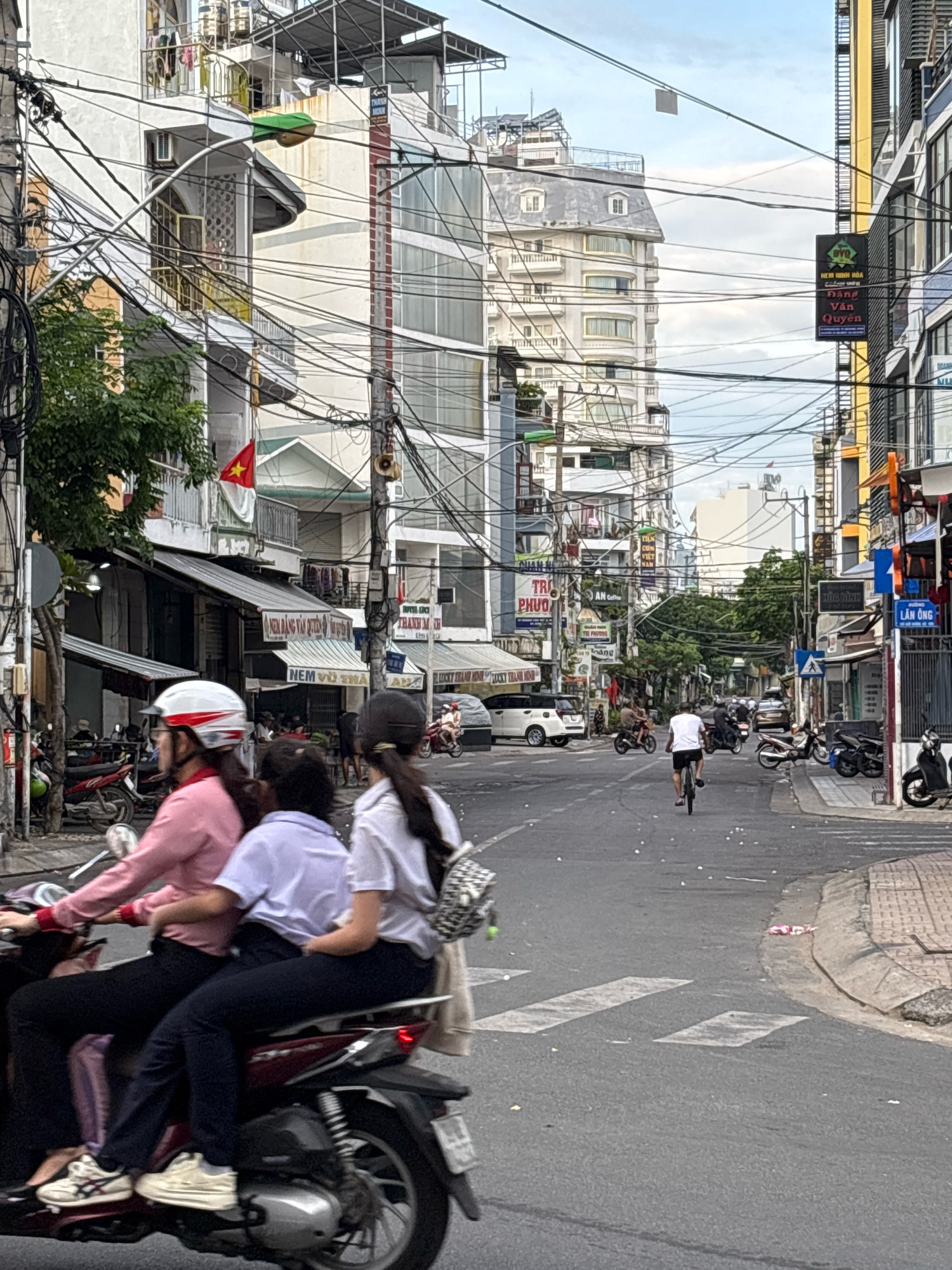 The image depicts a street scene in Nha Trang, Vietnam, characterized by dense urban development and a prevalence of overhead electrical wires. In the foreground, three individuals are riding on a single motorbike. The person in the front is wearing a pink long-sleeved shirt and black pants, with a white and red helmet. The middle passenger wears a white school uniform shirt and dark pants. The third passenger, at the rear, is wearing a white shirt. The motorbike is dark red.
The street is paved with asphalt and marked with pedestrian crossings. Various buildings line the street, displaying a mix of architectural styles, including multi-story residential and commercial structures. Many of these buildings feature balconies and awnings. Signs in Vietnamese are visible on the storefronts, indicating businesses. In the background, the sky is overcast. Other vehicles, including motorbikes and cars, are present on the street, suggesting typical urban traffic. A person is seen riding a bicycle further down the street. The overall impression is of a busy, developing city.