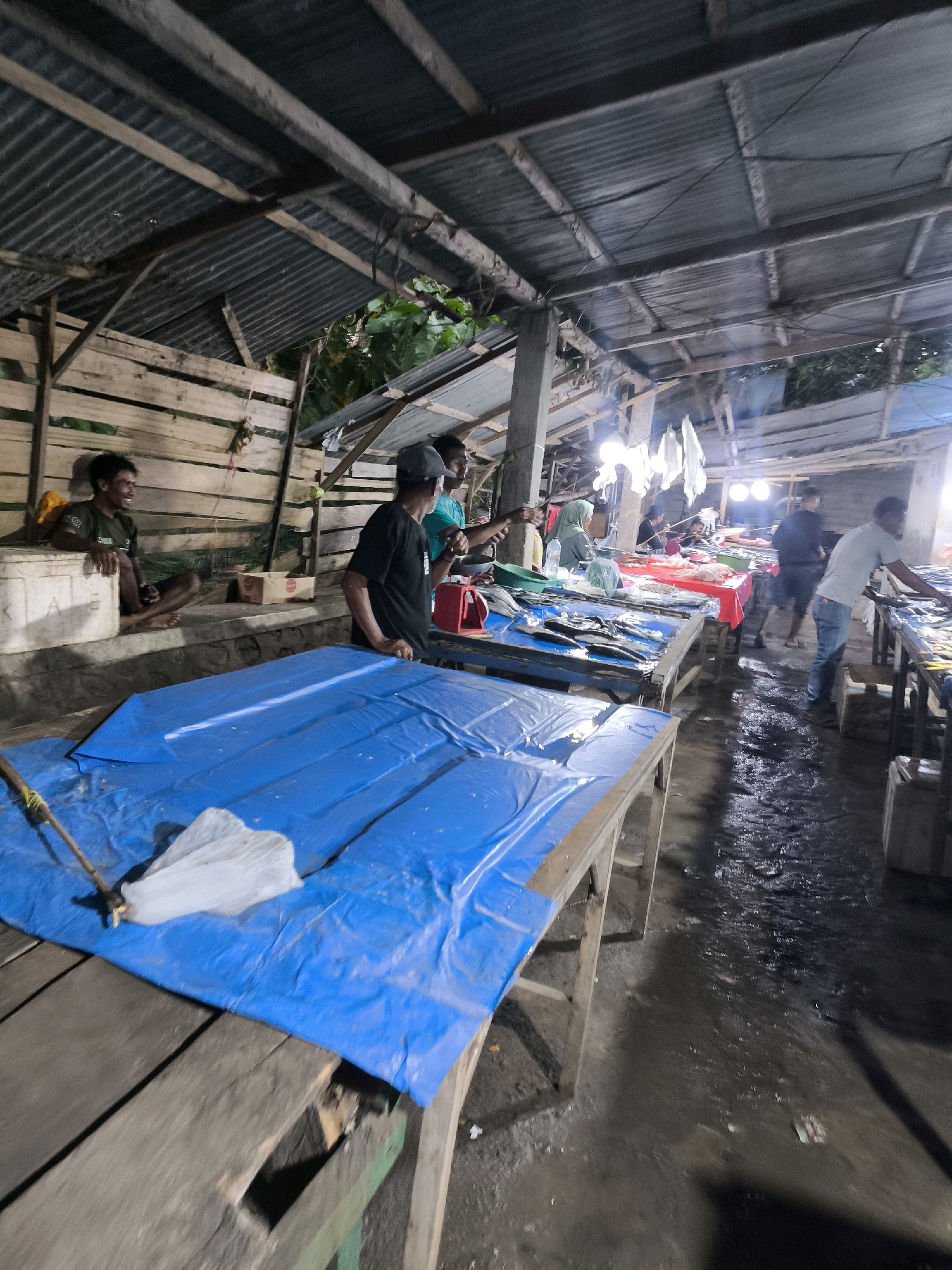 The image depicts a fish market scene, likely in a wet, outdoor or semi-enclosed area, possibly in Indonesia given the context. The overhead structure appears to be constructed of corrugated metal roofing supported by wooden beams and posts. The floor is wet and appears to be muddy or dirt, with water pooling in places.
In the foreground, a wooden table is covered with a bright blue tarp, on which a white plastic bag is placed. Another wooden table with a similar blue tarp is partially visible to the left.
In the mid-ground and background, several stalls are set up with people attending to them. One stall features tables covered in red and blue cloths, displaying numerous fish. People are seen browsing and possibly purchasing fish. There are also several individuals visible, some appearing to be vendors and others customers. One man in a green shirt is seated on a white cooler on the left side of the image. Another man wearing a black t-shirt and a baseball cap is standing and looking towards a stall. A woman in a hijab is also present, attending to a stall. The lighting in the market appears to be a mix of natural light filtering through the roof and artificial lights, including bright bulbs hanging from the ceiling, creating a somewhat dim and hazy atmosphere.
The overall impression is a bustling, functional marketplace where fresh fish is being sold.