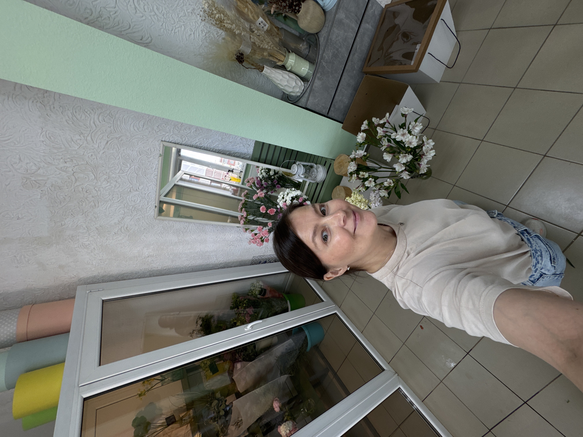 An indoor selfie, rotated 90 degrees clockwise, captures a woman with dark hair and a light-colored long-sleeve top, wearing denim bottoms, smiling subtly at the camera. Her face is in the upper right quadrant of the image.
The setting appears to be a flower or gift shop. The walls and ceiling are covered in white embossed wallpaper with a swirling pattern. A light green horizontal band separates parts of the wall. To the left and behind the woman, a mirror reflects an interior space and what seems to be a window looking out onto buildings.
Various floral arrangements and decorative items are visible throughout the space. Behind the woman, on a dark countertop or shelf, several vases hold dried flowers and grasses. Below the woman and slightly to her right, two glass-fronted display cases showcase bouquets of fresh flowers in different colors, including pink, white, and green. Below these cases, rolls of colorful packaging paper (pink, light blue, yellow, and light green) are stacked horizontally.
On the tiled floor, which is light beige with a grid pattern, are white gift bags with black handles and open cardboard boxes. Further back, near the mirror, a white decorative lantern and more fresh flowers, like white alstroemeria and pink blooms, are arranged. The lighting suggests it is daytime. No visible text is present.