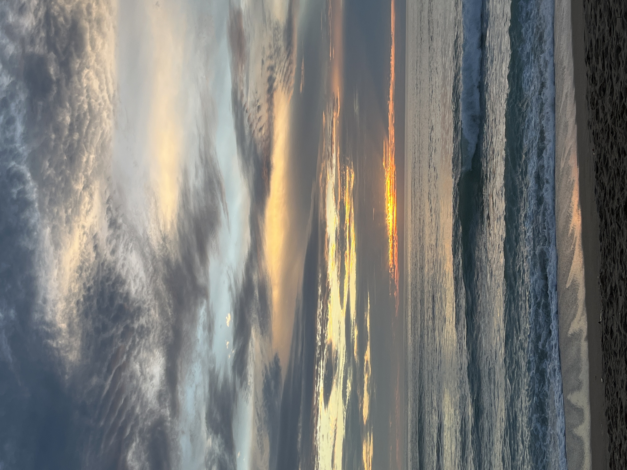 The image depicts a serene beach scene in Kamala, Thailand, likely during sunrise or sunset. The sky is filled with dramatic clouds, painted with hues of gray, blue, yellow, and orange, indicating the presence of the sun near the horizon. The sun itself is a bright, concentrated band of yellow and orange light peeking through the clouds just above the sea.
The sea is calm with gentle waves rolling towards the shore. The water displays various shades of blue and green, reflecting the colors of the sky. The waves create a foamy white crest as they approach the sandy beach.
The foreground consists of a stretch of dark sand, with some subtle texture indicating footprints or ripples. There are no people or artificial objects visible in the image, presenting a natural and untouched coastal landscape. The overall atmosphere is tranquil and picturesque.