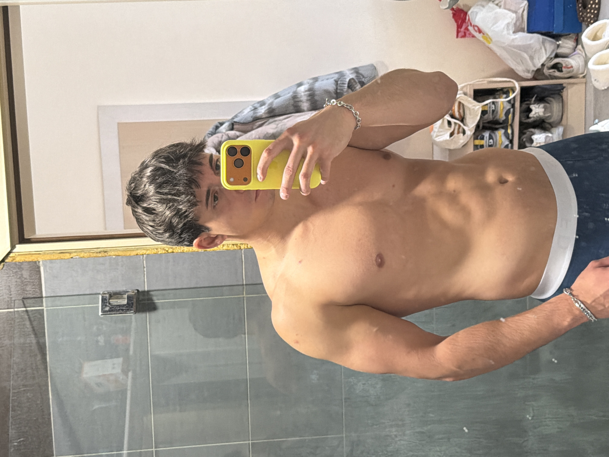 A young man is captured in a mirror selfie, shirtless and posing indoors. He has dark, short hair and is holding a bright yellow smartphone with a visible multi-lens camera array in his right hand, partially obscuring the lower half of his face. His visible left eye looks directly towards the camera with a neutral expression.
He exhibits a lean, somewhat muscular build, with visible abdominal definition. He is wearing dark blue underwear or swim trunks with a white waistband. A silver-colored chain bracelet is worn on his left wrist. Notably, there are several light-colored patches visible on his skin across his torso and arms.
The background, seen as a reflection in the mirror, reveals a domestic setting. A plain light-colored wall is visible, with a light blue and grey striped garment hanging in the upper left. A white-framed item, possibly a picture, is also reflected. To the right, a shelving unit stores several pairs of shoes, primarily sneakers, and various plastic bags (white, red, yellow). The immediate foreground shows grey tiled walls, suggesting a bathroom or a similar tiled area, with a small metal fixture on the wall. A window frame is partially visible along the left edge of the reflection. The lighting is bright and natural, indicating daytime. There is no visible text in the image. The scene conveys a personal moment in a home environment in Sofia, Bulgaria.