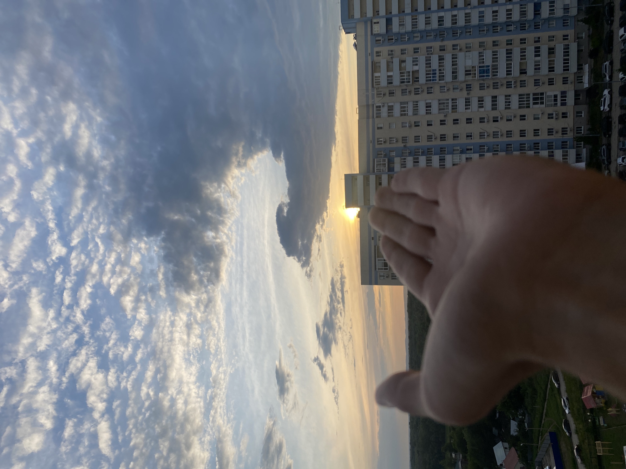 A first-person perspective shot captures a hand reaching out towards a sunset in Novokuybyshevsk, Russia. The sun, a bright orb partially obscured by clouds, is visible near the horizon, casting a warm glow across the sky. A tall, multi-story apartment building with light blue and yellow facades dominates the middle ground. Below the building, a dense line of trees marks the edge of a residential area with houses and a playground. Cars are parked along a road in the foreground. The sky is filled with a mix of fluffy white clouds and darker, more ominous gray clouds, creating a dramatic contrast with the warm hues of the sunset.