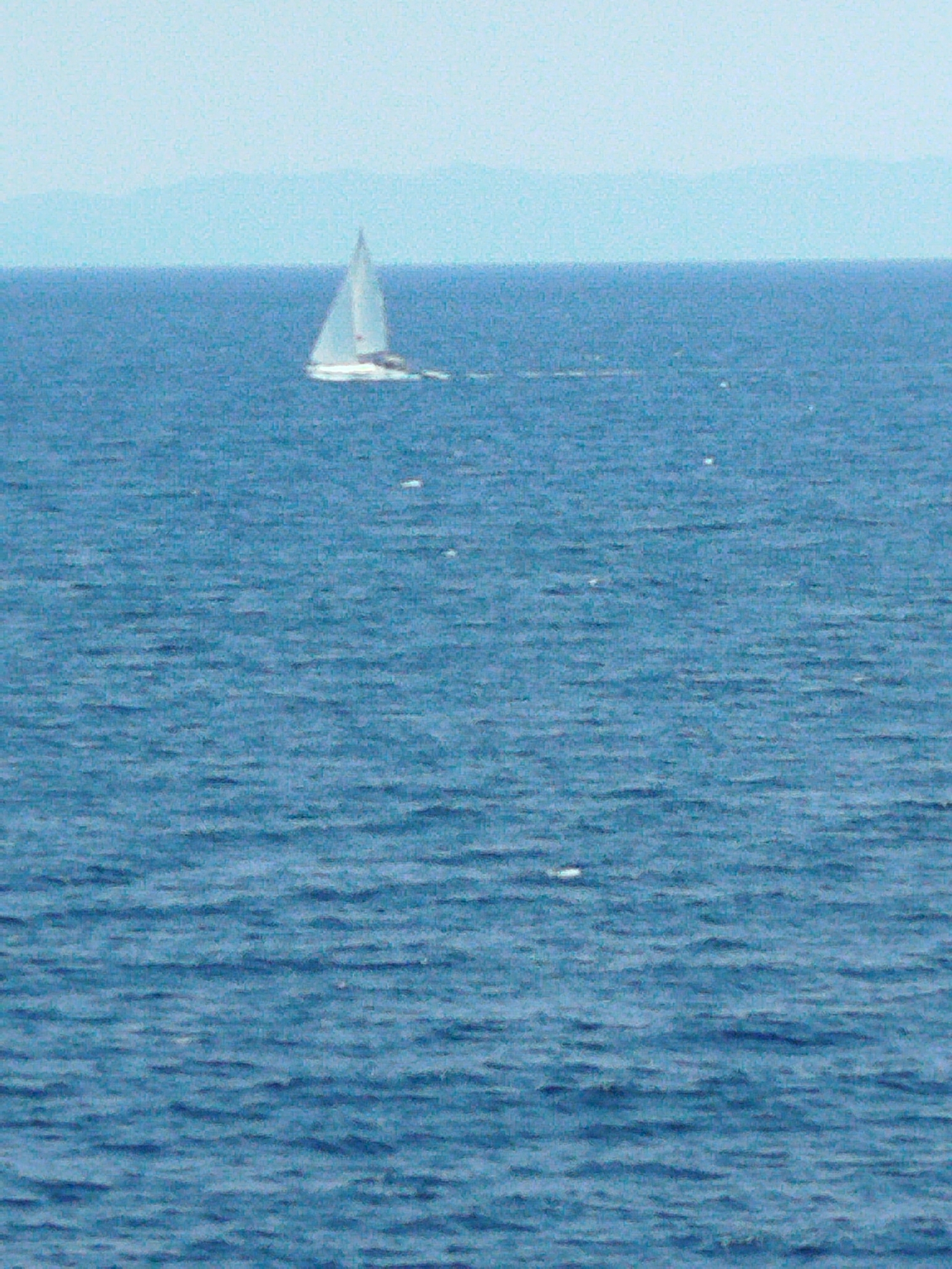 A lone sailboat with white sails navigates calm, blue waters under a hazy sky. In the distance, faint, layered mountain ranges are visible on the horizon, suggesting a coastal or island setting. The water is moderately choppy with small ripples. The scene appears serene and undisturbed. The image is set in the Sithonia Municipal Unit, Greece.
