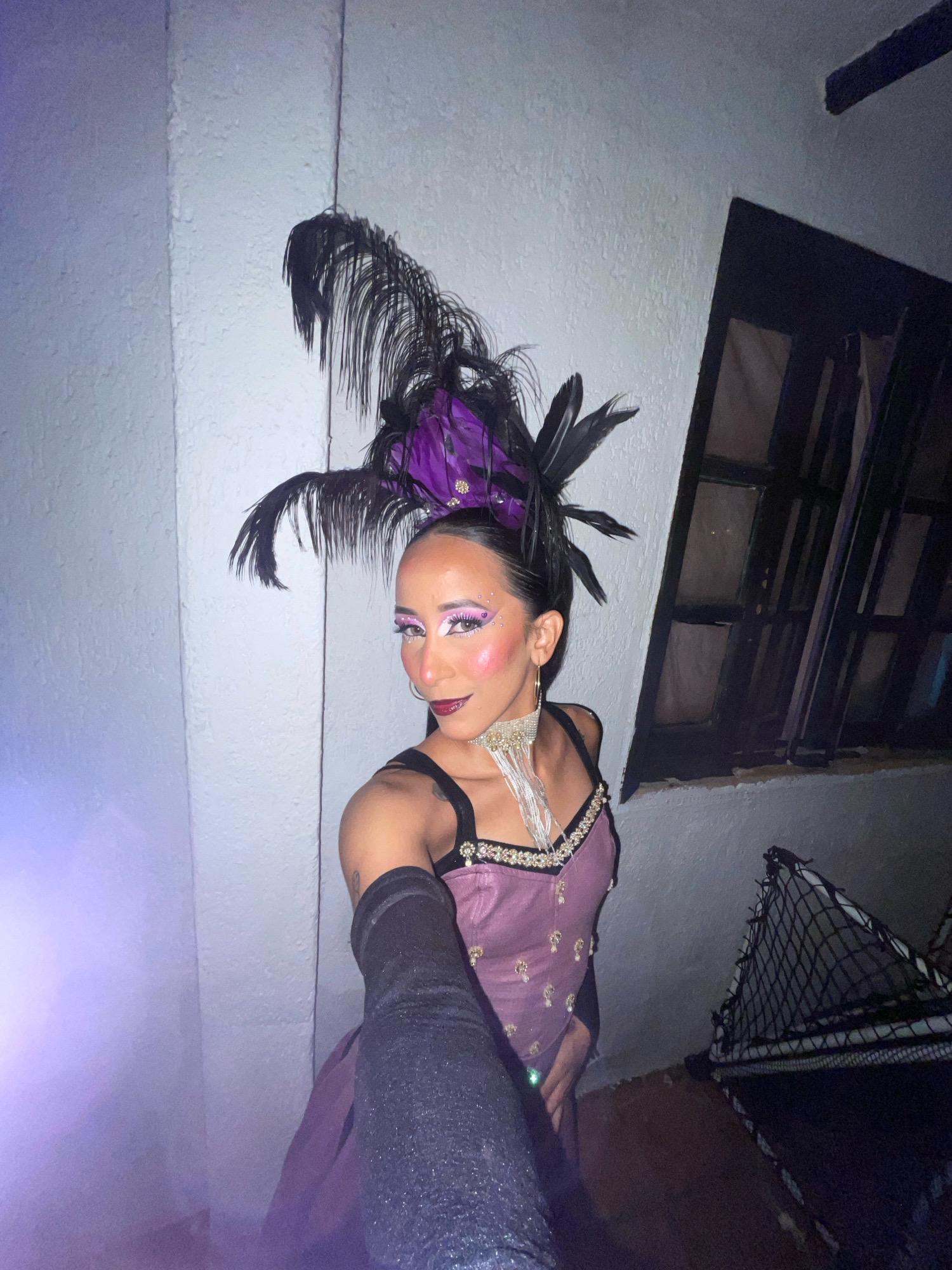 A person wearing a purple and black costume with a feathered headdress, heavy eye makeup, and a sequined necklace poses for a selfie in an alleyway. The person has dark hair pulled back and is wearing long black gloves. The background consists of a textured white wall on the left, a dark window with visible bars on the right, and a black net-like object on the ground in the lower right corner. The lighting is dim with a purple hue emanating from the left side of the frame. The person is standing and appears to be at an event or party. The location is specified as an alleyway in Porlamar, Venezuela.