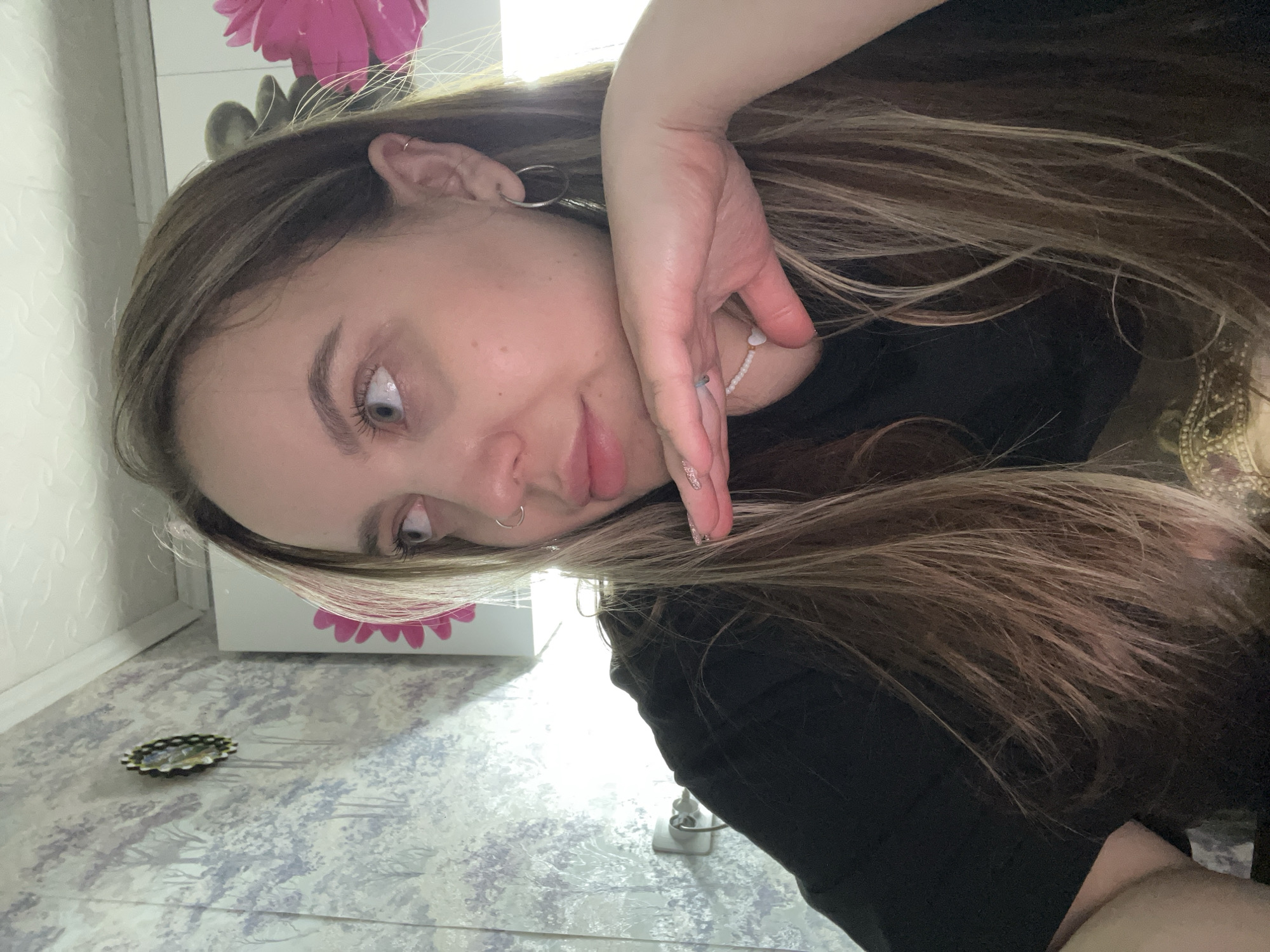 A young woman with light brown hair and blue eyes is posing for a selfie indoors. She is wearing a black shirt and has a silver hoop earring in her left ear, as well as a nose ring on her left nostril. Her right hand is positioned near her face. The background includes a wall with a floral design, a white cabinet or box with a pink flower decal, and a tiled floor with a patterned design. An ornamental object is on the floor in the lower-left corner. A power outlet and cable are visible behind her torso. The image was taken with a tilted perspective. There is no verifiable evidence that connects the image to Novopokrovskaya, Russia.