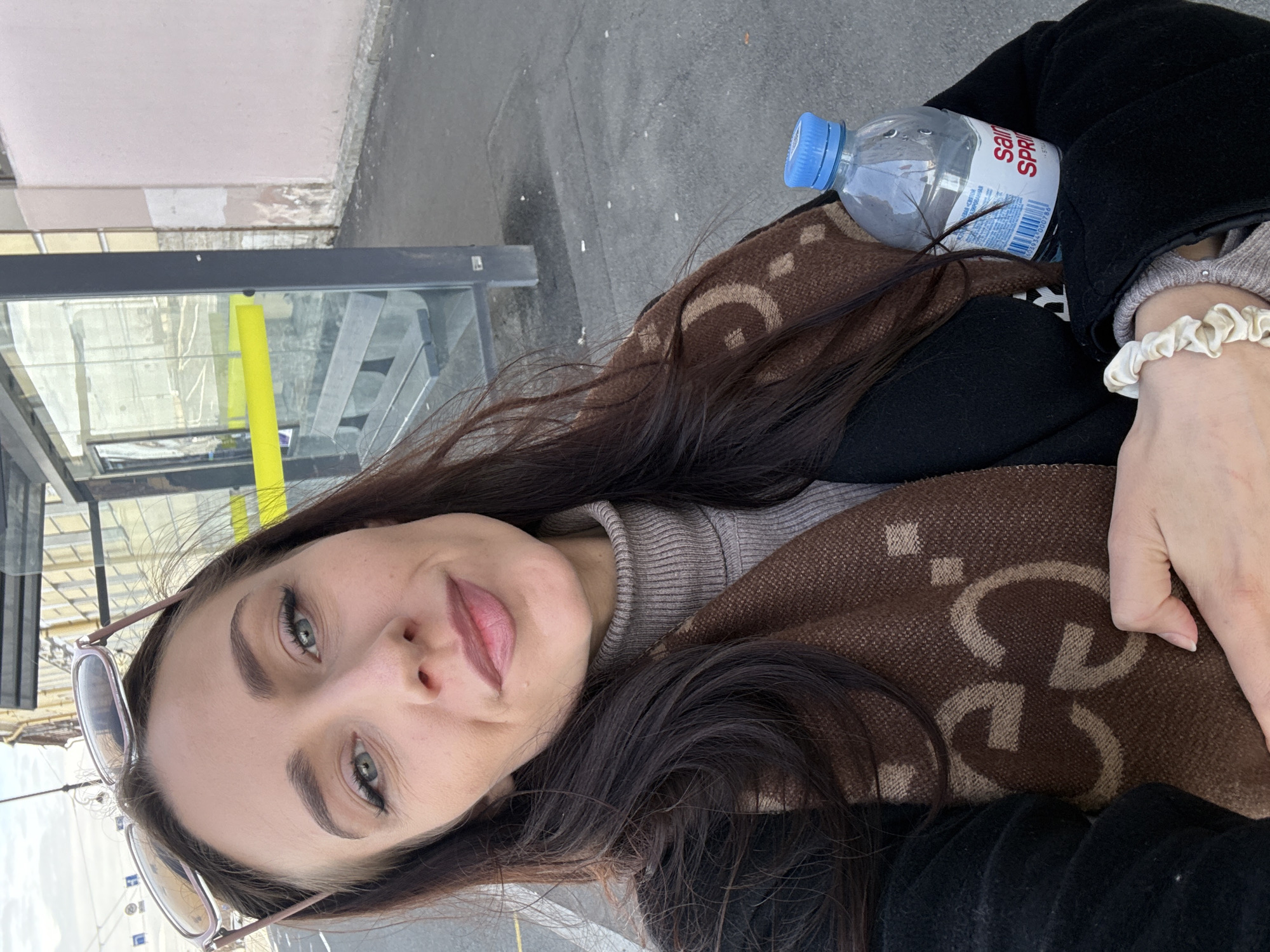 A young woman is featured in a close-up, chest-up selfie, taken outdoors in what appears to be an urban setting in Saint Petersburg, Russia. She has long, dark brown hair and looks directly at the camera with a slight, relaxed smile. Her eyes are green, enhanced with eyeliner, and her eyebrows are well-defined. Sunglasses are perched on top of her head.
She is wearing a light grey turtleneck sweater underneath a black coat. A brown scarf with a prominent repeating interlocking "GG" pattern is draped around her, indicative of the Gucci brand. On her right wrist, a light, possibly cream-colored, ruffled scrunchie is visible.
Next to her, a clear plastic water bottle with a blue cap is present. Part of the label on the bottle reads "SAINT SPRING." The background includes a glass-paneled structure, likely a bus stop or shelter, behind which a bright yellow vertical bar is visible. The ground is paved, resembling asphalt. The scene is well-lit by natural light, suggesting it is daytime, portraying a casual moment in a city environment.