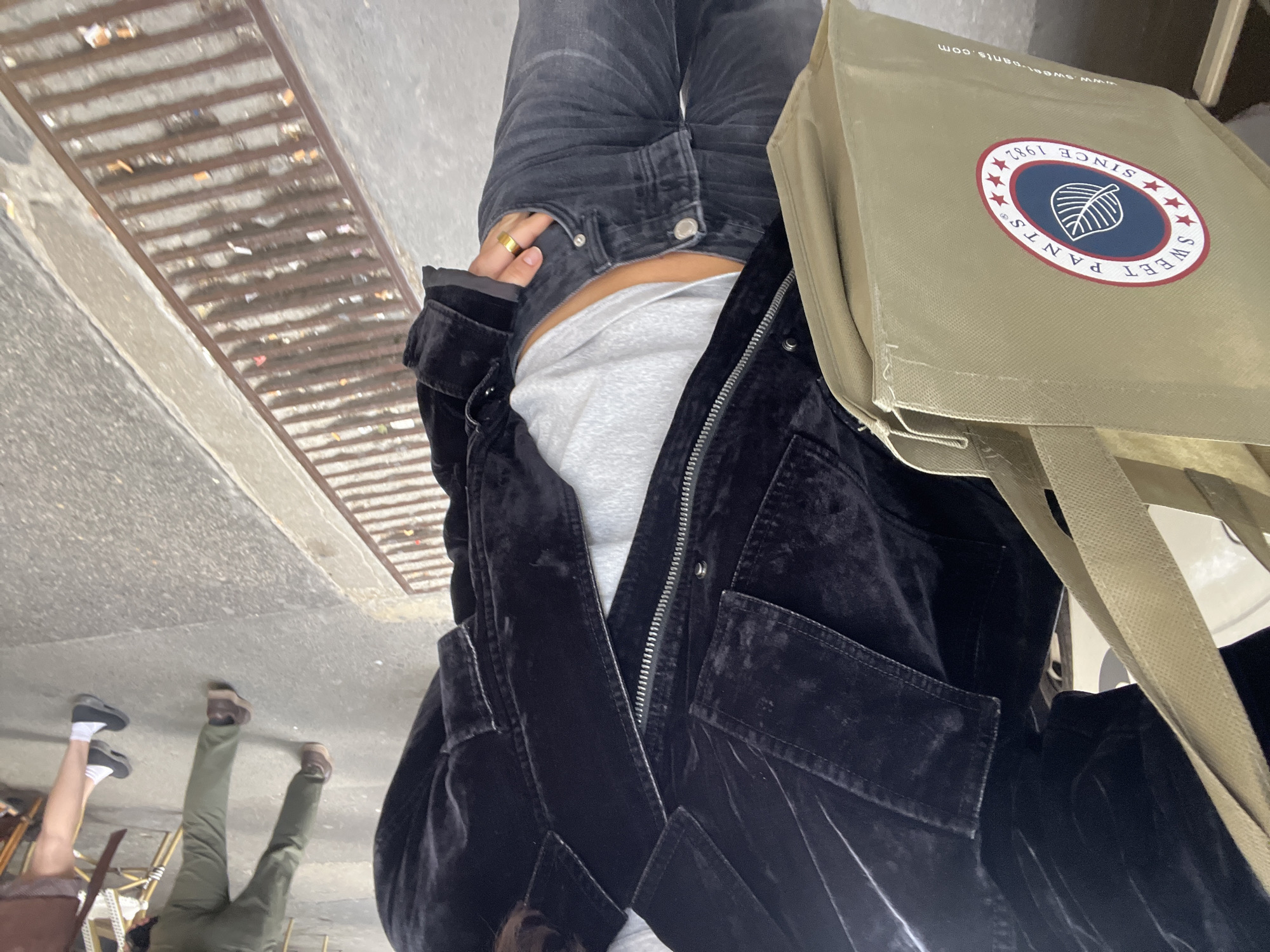 The image, taken from a slightly downward, first-person perspective, depicts a person standing or walking on an urban sidewalk. The main subject wears a black velvet jacket, a grey undershirt, and dark grey jeans, with a gold ring visible on their finger. They carry a light olive green canvas tote bag.
The setting is an outdoor city environment, likely a street or pedestrian area in Paris, France. The foreground features concrete or asphalt pavement and a long metal grate, possibly a storm drain, which contains visible debris such as cigarette butts and small pieces of paper.
In the background, two other individuals are visible from the waist down, appearing to be seated at outdoor tables. One person wears green trousers and brown shoes, while the other shows bare legs with white socks and dark shoes. Legs of chairs are also discernible around them.
The activity suggests the main subject is in motion or paused on the street, while the background figures are relaxing at what appears to be an outdoor cafe.
Notable details include the diffused daylight, implying an overcast day or shade. The canvas tote bag prominently displays a logo with the text "SWEET PANTS" and "SINCE 1982" encircling a leaf motif. Part of a website address, "www.sweet-pants.com", is also visible on the bag. Emotions are not discernible from this perspective.