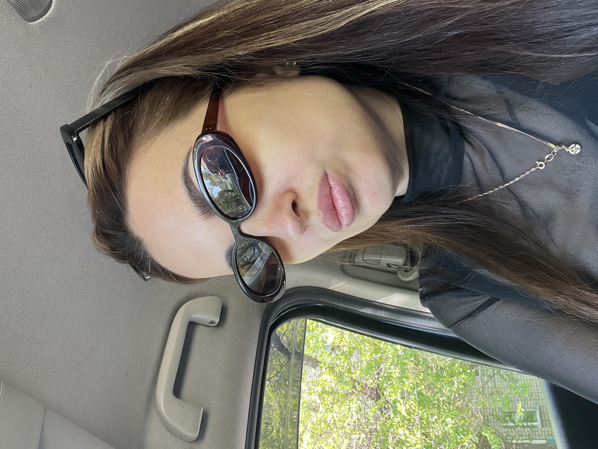 A woman is sitting in a car, wearing sunglasses and a black, mesh turtleneck. She is wearing a thin gold necklace. The car window shows trees and a building. The car's interior is gray.