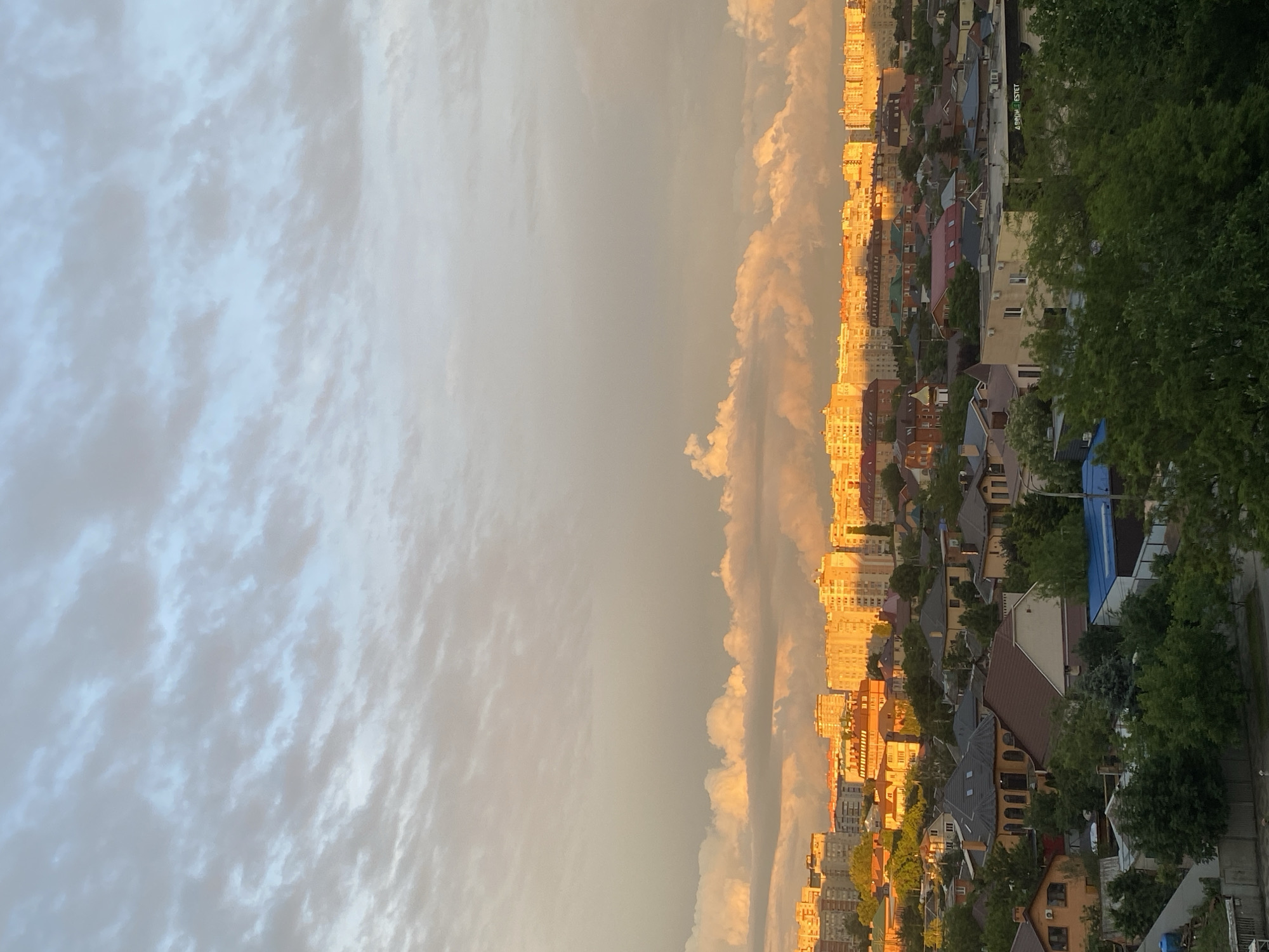 The image presents a panoramic view of a cityscape under a dramatic sky, captured during what appears to be either sunrise or sunset. The scene is dominated by a clear horizon line, with the sky occupying the upper half and urban development filling the lower half.
The sky on the left side is covered by extensive, lighter grey and white clouds, suggesting a partially overcast day. Towards the center and right, the clouds take on warm, golden-orange hues, intensely illuminated by the low-angled sun. This golden light also dramatically bathes the buildings along the horizon.
The cityscape below consists of a mix of structures. On the left, a dense cluster of multi-story buildings, likely apartment blocks, glow with the golden light, creating a striking contrast against the muted sky. Moving towards the right, the buildings become less uniform, transitioning into a mix of houses and smaller residential structures with varied roof colors and designs. Many of these houses are partially obscured by lush green trees, particularly on the far right side, which suggests a greener, possibly more suburban or park-like area bordering the city. No people or specific activities are visible within the frame.
A notable detail is some text visible on a building in the mid-ground on the right side, which reads "APARTMENT ESTET". The overall mood conveyed by the lighting is serene and picturesque, highlighting the beauty of the "golden hour."