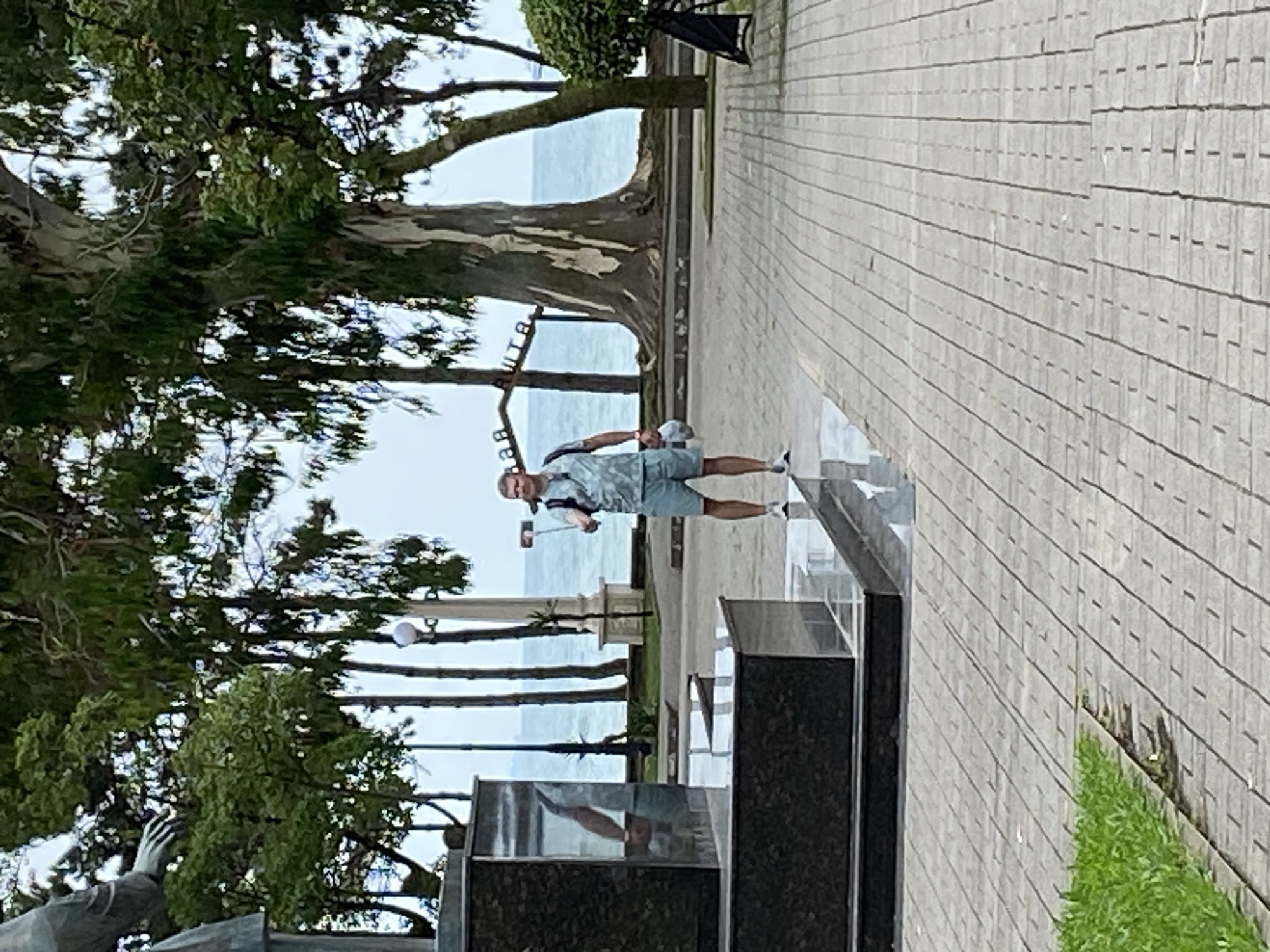 A man is standing on a paved walkway in front of a large monument, holding a selfie stick with a phone attached. He is wearing a light-colored patterned short-sleeved shirt and light-colored shorts. He has a backpack on and is holding a white hat in his left hand. In the background, there is the sea and a large tree. The location is Sokhumi, Abkhazia.