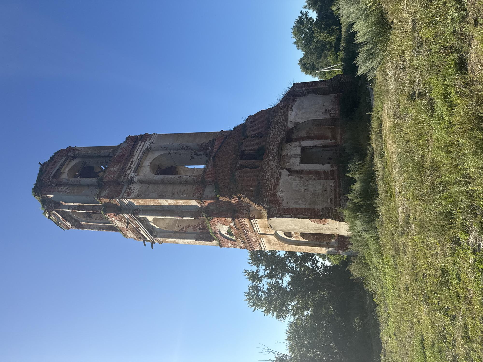 An old, dilapidated bell tower stands tall against a clear blue sky. The tower is made of brick and stone, with visible signs of decay and wear. Arched openings are visible at various levels, some of which are boarded up or broken. The top of the tower is crumbling, with vegetation growing from the cracks. The tower is surrounded by overgrown grass and some trees on either side. The sun is shining brightly, casting a slight glare on the left side of the image. The scene appears to be in a rural or abandoned area.