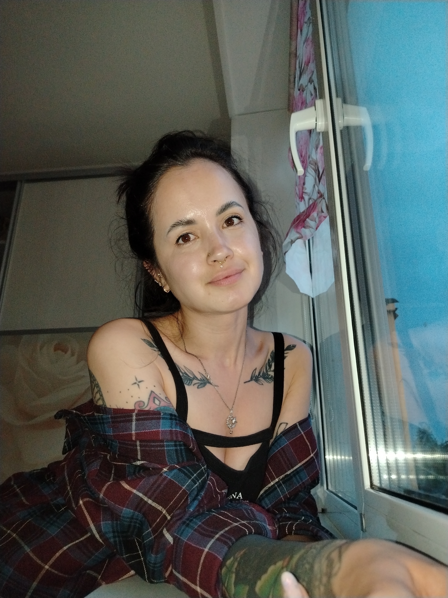 A woman with dark hair pulled back in a messy bun is pictured indoors next to a window. She has a nose ring and is wearing a black tank top and a plaid shirt draped over her shoulders. Tattoos are visible on her arms and chest. The window reflects an outside view, possibly at twilight or dusk. The environment appears to be a room with a window frame and curtain visible. The image provides no information to confirm the city of Noviy, Russia.