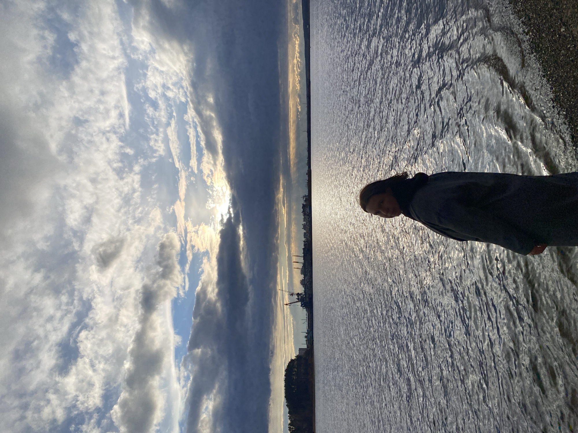 The image, correctly oriented, displays a wide landscape featuring a large body of water under a dramatic sky. The scene is captured during what appears to be late afternoon or early evening, given the low sun and the golden light reflecting off the water.
In the foreground, on the right side of the frame, a person is partially visible from the chest up, looking downwards and slightly to the right. They have dark hair and are wearing a dark hooded garment. The shoreline immediately adjacent to them is dark and appears rocky or pebbly where the water meets the land.
The vast expanse of water dominates the right two-thirds of the image, shimmering brightly with reflections of the sun, creating a textured surface of light and shadow. Across the water, on the distant horizon, a long, low strip of land is visible, featuring silhouettes of industrial structures, including what appear to be cranes and buildings, suggesting a port or industrial area.
The sky occupies the left third of the image and is filled with a dynamic cloudscape. White, wispy cirrus clouds mix with larger, darker cumulus or stratocumulus formations. Patches of clear blue sky are visible between the clouds, and sunlight breaks through the cloud cover on the left, casting a bright glow.
No specific activity is taking place beyond the person's presence by the water. No visible text is present in the image. Given the context of Chaikovsky, Russia, the water body is likely the Kama River or Votkinsk Reservoir.