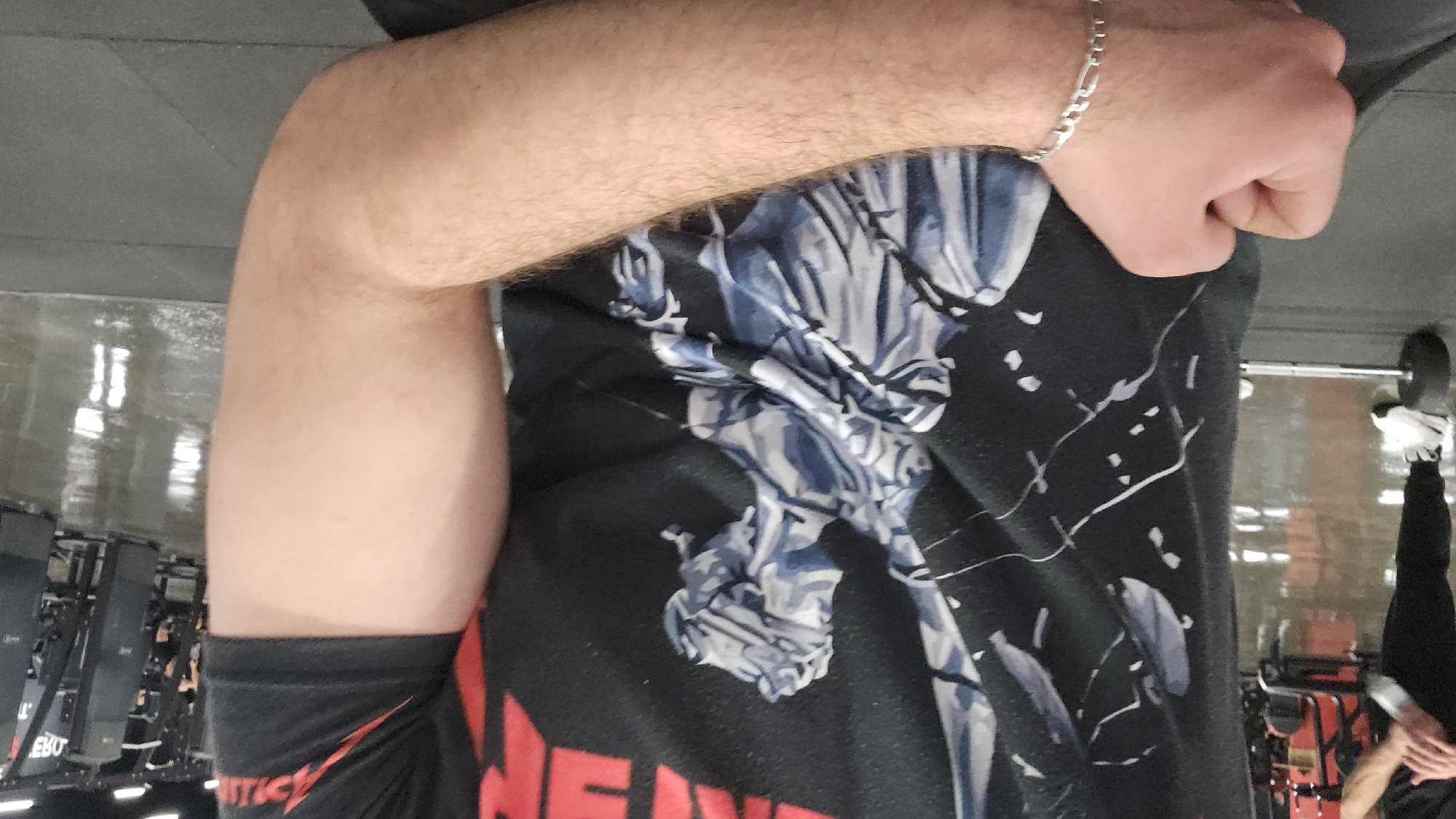 This image, taken in Plovdiv, Bulgaria, shows a close-up, first-person perspective of a person's upper body within an indoor gym setting. The primary focus is on the individual's right arm, which is flexed, revealing muscle definition, and their right hand, clenched into a fist. A silver figaro chain bracelet is worn on the person's right wrist. The skin tone is light, with visible arm hair.
The person is wearing a black short-sleeved t-shirt featuring a detailed blue and grey graphic design on the front, which appears to depict a stylized figure or abstract art. Prominent red text, spelling "NEVER," is visible at the bottom left of the shirt. Additional red lettering, possibly part of a word like "TICE," can be seen on the black sleeve.
The background reveals a dark, highly reflective floor, typical of a gym, showing streaks of light and clear reflections of equipment. On the left, several exercise machines, possibly rowing machines, are visible, with "EBO" partially legible on one. On the right, the reflection of another person's lower legs, clad in black pants and white athletic shoes, is visible on the floor, indicating the presence of others in the facility. The artificial lighting is consistent with an indoor environment. The scene suggests a moment either during or immediately after a workout, with the person seemingly flexing their arm.