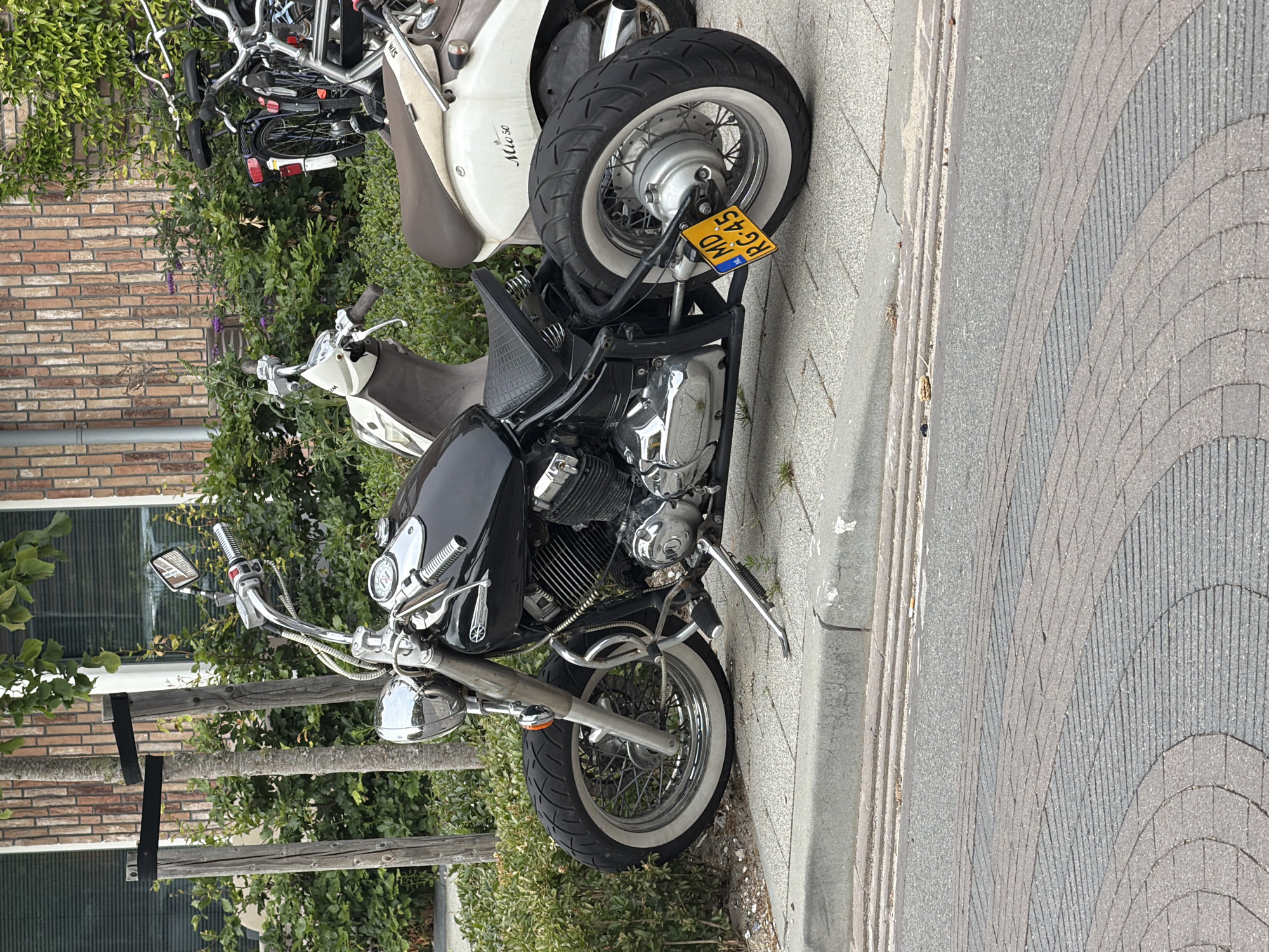 The image shows a black custom motorcycle parked on a sidewalk in Rijswijk, Netherlands. The motorcycle is positioned on the left side of the frame, with its front wheel slightly turned to the left. It has chrome accents, spoked wheels with white-wall tires, and a textured black seat. A yellow license plate with the inscription "MD RG-45" is attached to the rear fender. Behind the motorcycle, there are two scooters and some bicycles parked. The background consists of a brick building and lush green foliage. The ground is paved with interlocking bricks and asphalt.