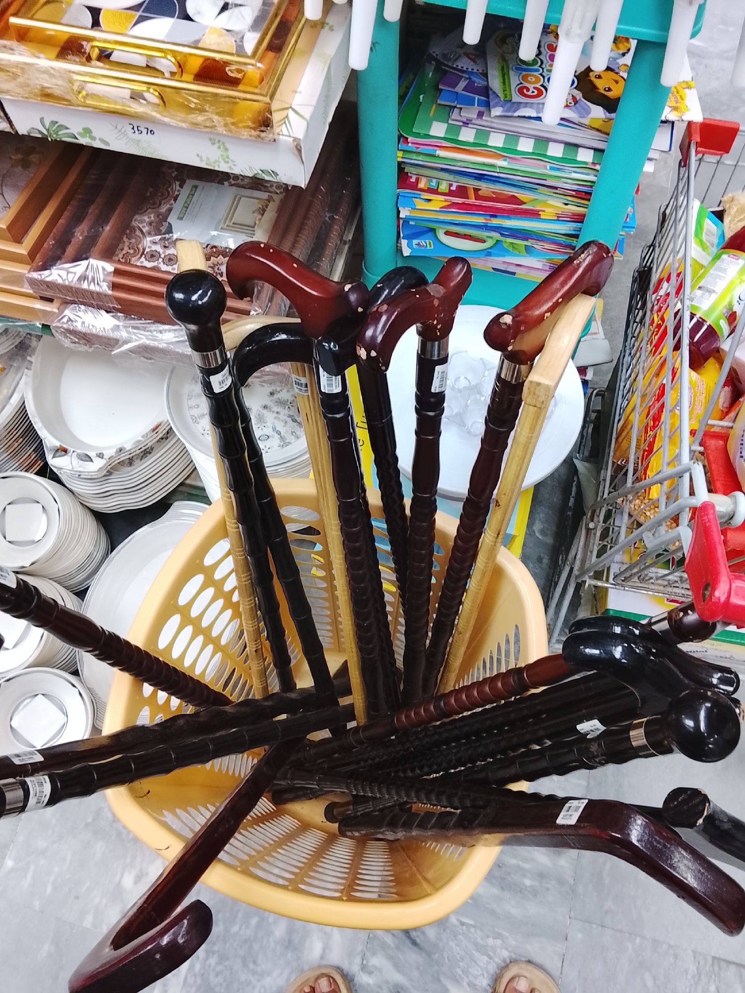 A high-angle, indoor shot depicts a collection of walking sticks arranged in a yellow, plastic, perforated basket. The sticks are made of dark and light brown wood, and vary in design, with some having rounded knobs and others having curved handles. A stack of white plates is visible to the left of the basket. In the background, partially obscured, are stacks of packaged items on shelves. To the right, a shopping cart contains food items. The environment suggests a retail setting. Partial view of feet (human) visible at the bottom of the frame. The location, Gulgasht Colony, Pakistan, is specified.
