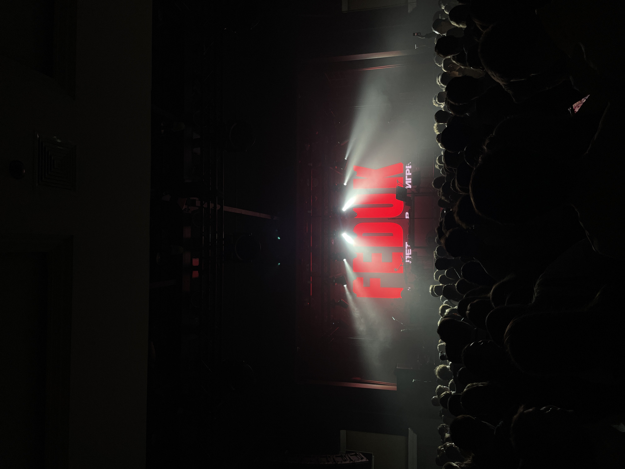 A concert is taking place in Kazan, Russia. The audience is closely packed on the right side of the frame, their heads silhouetted against the stage lights. Onstage, the word "FEED" is displayed in large, illuminated red letters, lit by white spotlights. The scene is dark, with stage lights creating a hazy atmosphere. The left side of the image features a wall with architectural details.