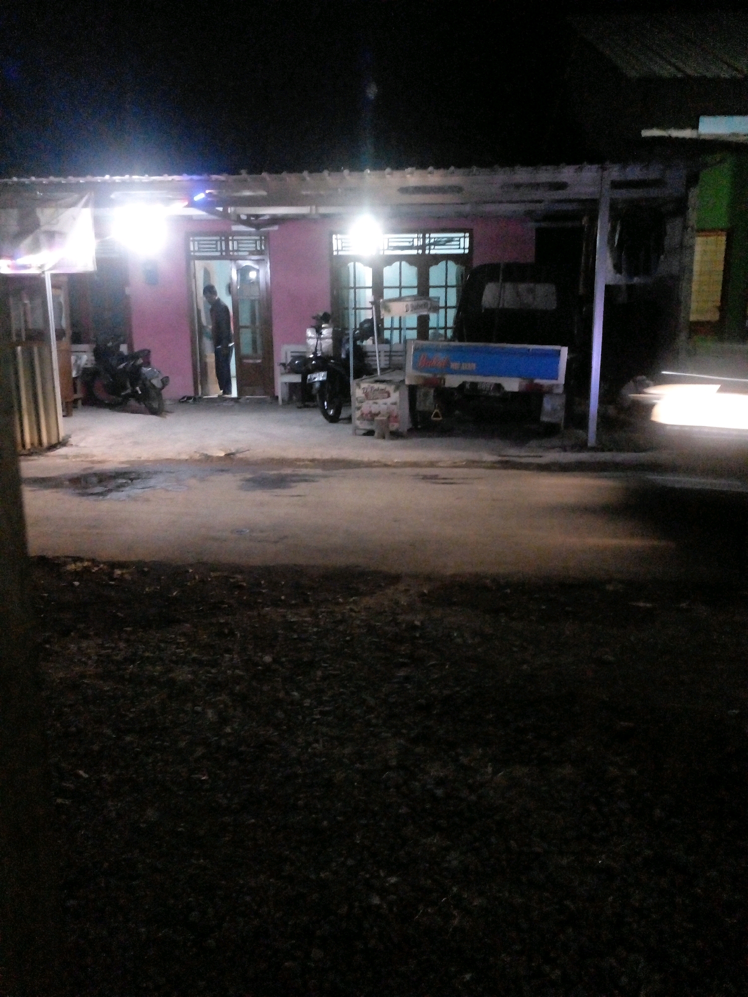 A nighttime scene in Pungangan, Indonesia, captures a street in front of a building with a pink facade. A person stands in the doorway of the building, illuminated by bright lights. To the left of the building, a motorcycle is parked. To the right, a small truck with a blue cargo bed is visible, along with another motorcycle. The ground in front of the building appears to be paved and wet, with puddles reflecting the light. The area is dimly lit, with the primary light sources being the building's interior and exterior lights. The overall impression is of a residential or small business area at night.