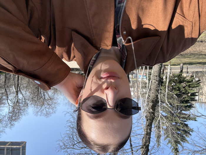 A woman is photographed outdoors in a close-up, selfie-style shot on a sunny day. She has dark hair pulled back and wears dark-tinted, angular sunglasses with a silver chain attached to the right earpiece. She is dressed in a collared jacket made of brown, suede-like material, worn over a visible plaid shirt and an underlying black garment. Her right hand is positioned near the back of her head and right ear. The background consists of a park-like environment with bare deciduous trees, scattered evergreen trees, and a paved ground surface. Multi-story residential buildings are visible in the far left background, partially obscured by bare trees and a metal fence. The location is identified as a park or public green space within Donetsk, Ukraine.