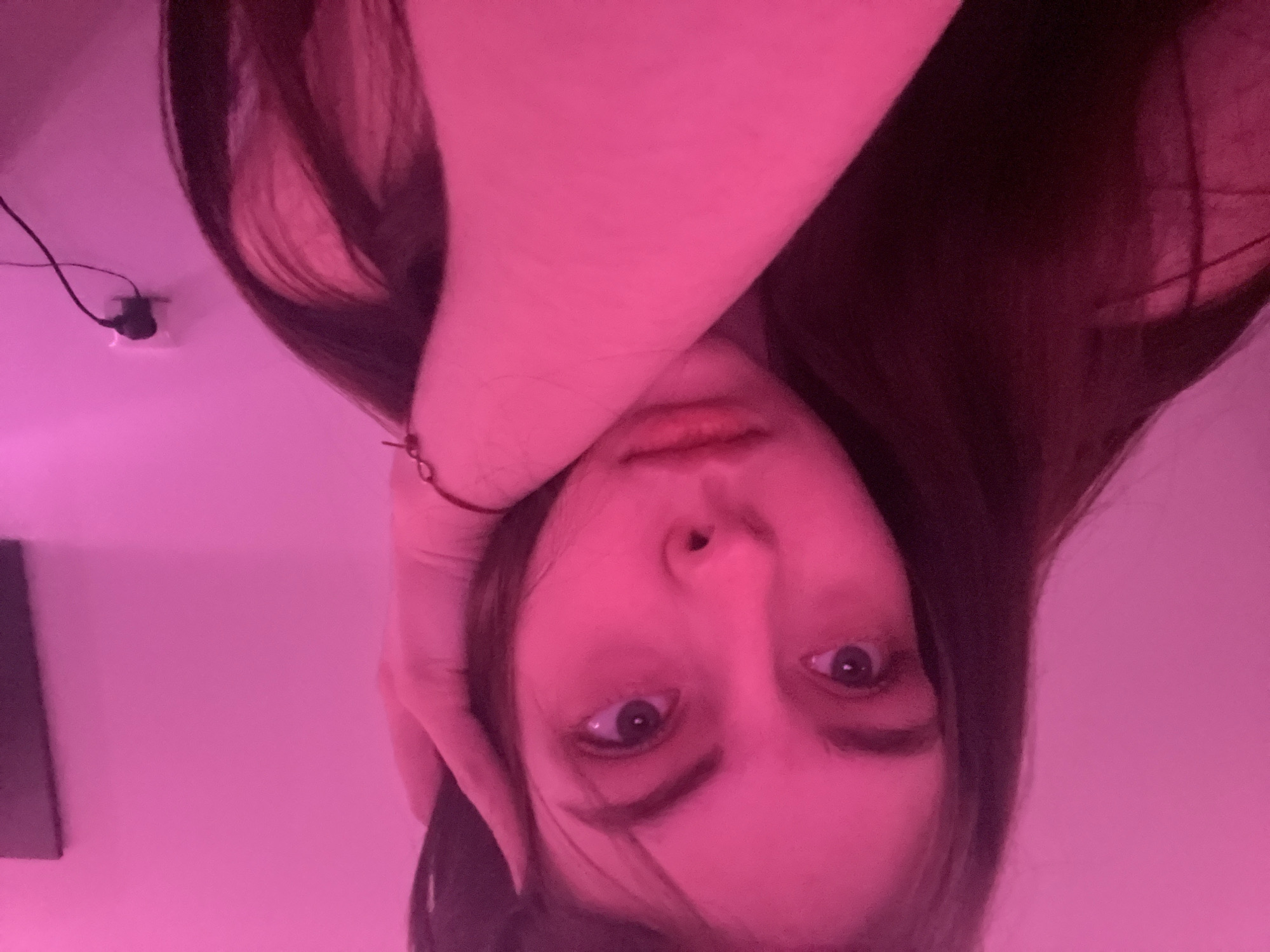 A female is facing the camera, with her head tilted downward. Her right hand is placed on the right side of her head. The environment is lit with a pink light. An electrical outlet and wire are visible in the upper left corner of the frame. A dark-colored object is in the lower left corner. The location is unknown.