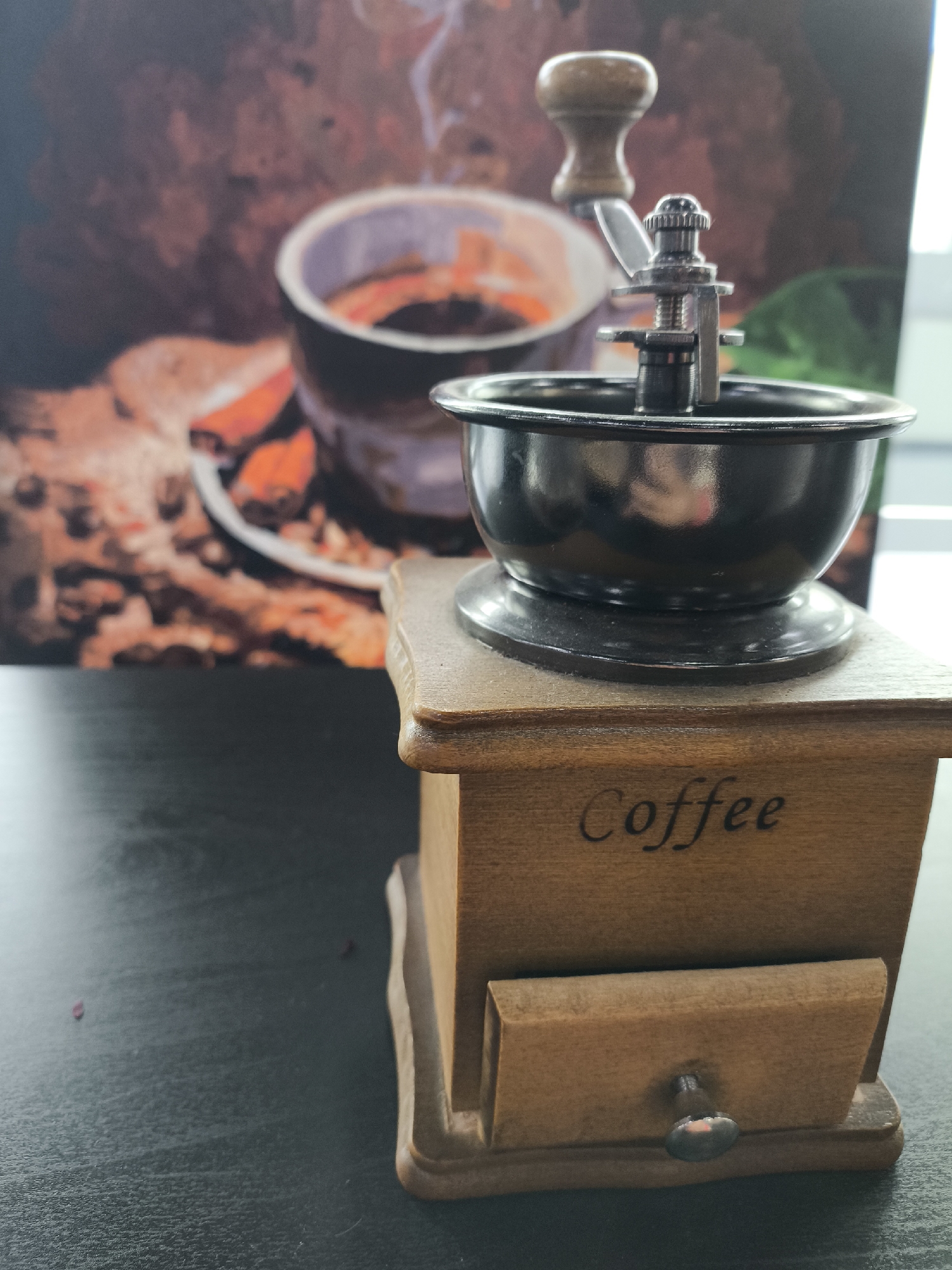 A coffee grinder sits on a dark, flat surface. The grinder is made of wood and has a small drawer at the bottom with a metal knob. The word "Coffee" is printed on the front of the grinder. Atop the wooden base is a metal bowl and grinding mechanism with a wooden handle. In the background, a blurred image shows a cup of coffee and pastries. The location is Unknown, Unknown.