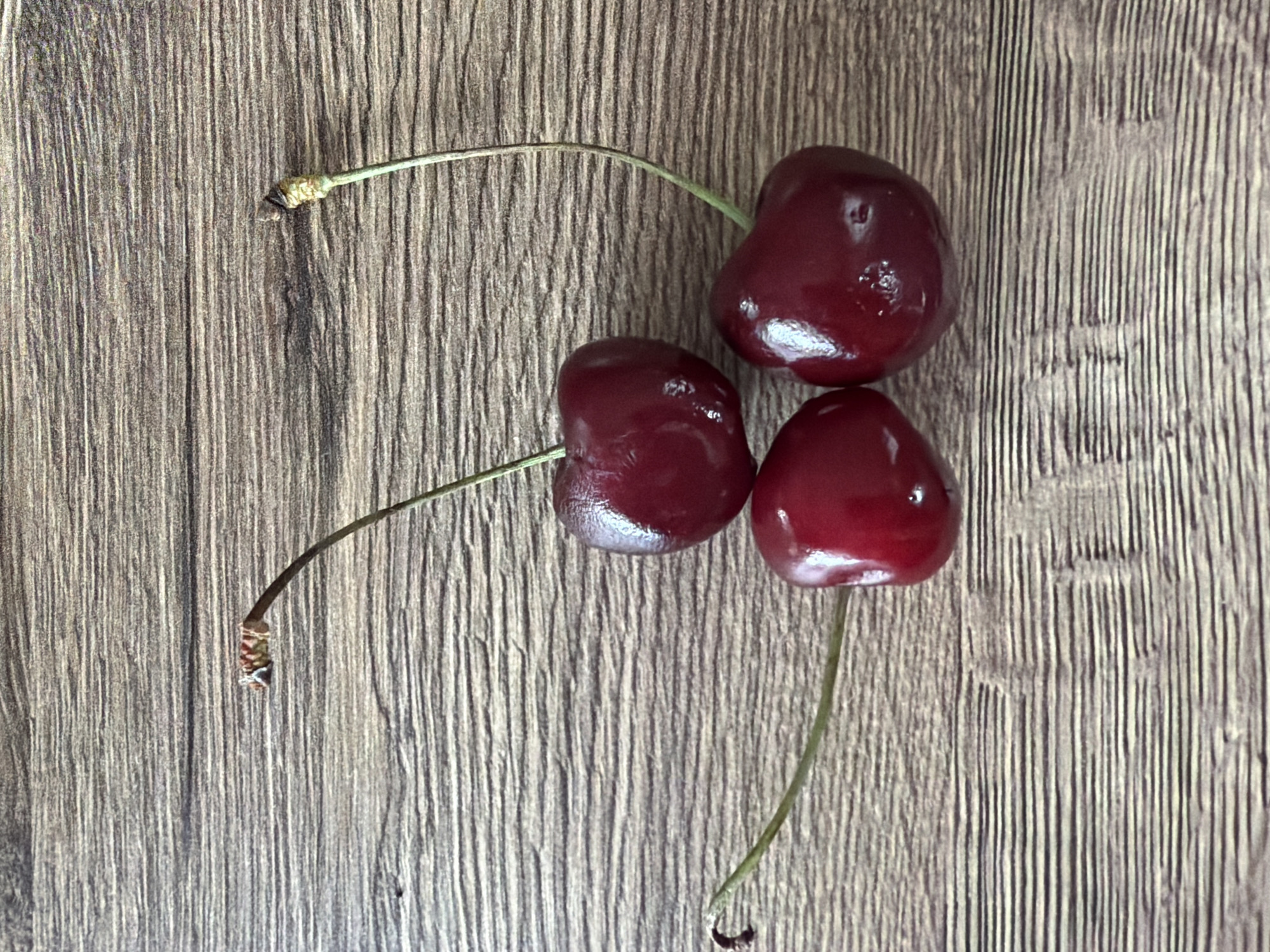 Three dark red cherries with thin green stems are arranged on a wooden surface with horizontal grain. One cherry is positioned at the top center, with its stem arching to the left. Another cherry is located below and to the left of the first, with its stem extending to the lower left. The third cherry is to the right and slightly below the first, with its stem curving upwards to the top right. The wooden surface is textured and appears to be a table or countertop. There are no people or animals present in the image, and no actions or interactions are depicted. The environment is an indoor setting, with artificial lighting. The location of Shashmurka, Russia is not depicted or implied by any elements within the image.