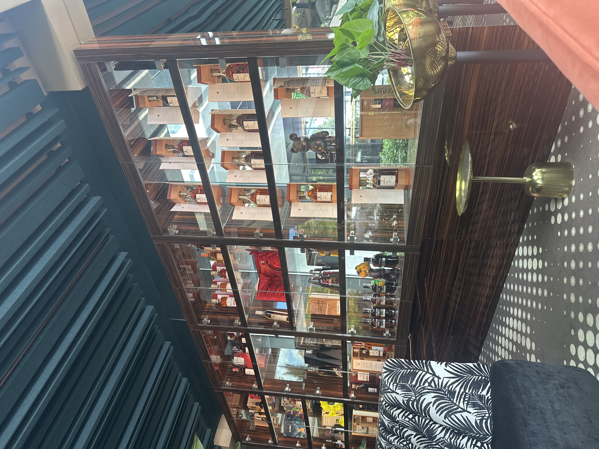 The image depicts an indoor bar area with a large, glass-fronted display cabinet filled with bottles of alcohol. The cabinet is well-lit, showcasing various brands and sizes of bottles, many in their original packaging. The bottles are arranged on multiple shelves, and the cabinet itself has a dark wood finish. In the foreground, a portion of a black armchair with a black and white patterned upholstery is visible. To the right of the armchair, a small, gold side table with a round top stands on a dark, patterned carpet. Further to the right, a large, gold planter holding a green plant is supported by a dark stand. In the background, beyond the display cabinet, a lush green outdoor space with trees and foliage is visible, suggesting the bar may have an outdoor seating area or a view of a garden. The ceiling above the display cabinet is constructed with dark, slatted panels. The overall ambiance appears to be sophisticated and luxurious.