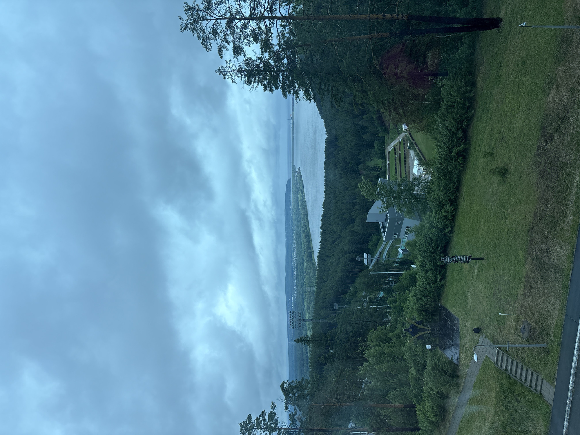 This image shows a view from an elevated position in Прикамский, Russia. The foreground features trees and vegetation, with a clear view of a river or lake in the distance. The sky is overcast with heavy clouds. The surrounding landscape includes forested areas and some signs of civilization.