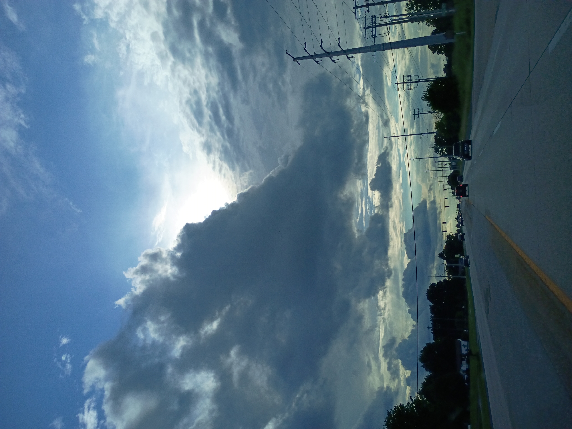 A wide-angle, low-angle exterior shot captures a highway scene under a dramatic sky. Dark, heavy clouds dominate the upper half of the frame, with patches of bright sunlight breaking through, creating a high contrast effect. The highway stretches forward, a grey asphalt road with a yellow line dividing lanes. Several vehicles are visible on the road, moving away from the viewer. Telephone poles with numerous wires line the right side of the highway, interspersed with trees and a few buildings visible in the distance. The overall impression is of a potentially stormy or overcast day.