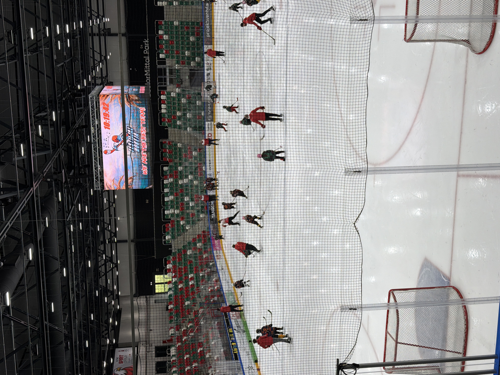 An aerial, wide-angle shot captures an ice hockey rink during what appears to be a training session. The rink is full of young players wearing various colored hockey uniforms, predominantly red, yellow, and black. Several adults, likely coaches, are also present on the ice, some of whom are wearing red jackets. The players are spread across the ice, engaged in drills, with hockey sticks in hand and pucks visible. Two hockey nets are positioned at either end of the rink. The stands, filled with red and green seats, are visible in the background, mostly empty. A large video screen hangs above the rink, displaying graphics and the time "10:19:42". The overall environment is that of an indoor sports arena, with exposed ceiling infrastructure and bright lighting. The location is the Arena Sosnowiec, located in Sosnowiec, Poland, a multi-purpose indoor arena.