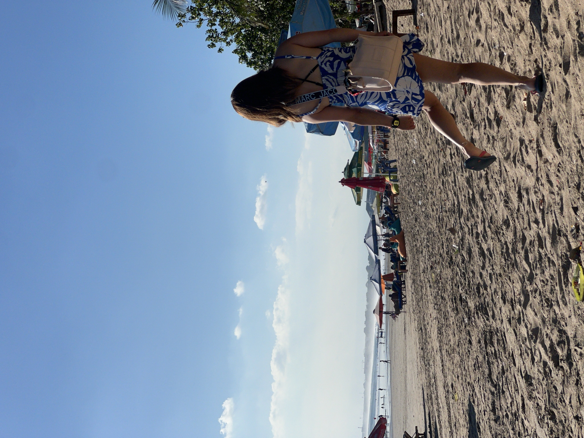 This media file depicts a sunny day at a beach in Kuta, Indonesia. The main subject is a woman with long brown hair, wearing a blue and white patterned dress and a light-colored shoulder bag. She is standing on the sandy beach, facing away from the camera, with her right leg bent. Her sandal is visible on her right foot, and she is wearing a watch on her left wrist.
In the background, there are several beach umbrellas, some open and providing shade, others closed. People can be seen sitting at tables and chairs under the umbrellas, enjoying the beach atmosphere. Further in the distance, the ocean stretches out, and there are faint outlines of buildings or structures along the coastline. The sky is a clear blue with a few scattered white clouds. The overall scene suggests a relaxed and pleasant day at the beach.
The text "MARC JACOBS" is visible on a strap of the woman's bag. The lighting indicates it is daytime, likely late morning or early afternoon, given the bright sunlight casting shadows. The presence of umbrellas and people enjoying the beach suggests a tourist-friendly location.