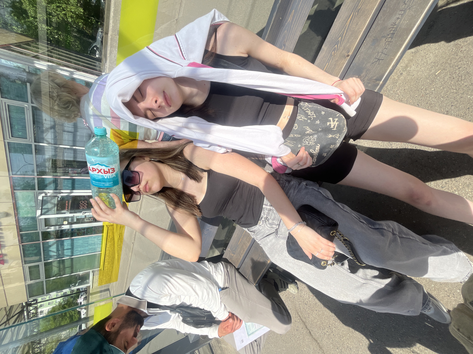 Two young women are sitting on a wooden bench in an outdoor urban setting. One woman wears a black tank top and black shorts, with a white sweatshirt draped over her head and shoulders. She holds a gray patterned purse. The other woman wears a black tank top, gray jeans, and sunglasses. She is holding a clear plastic water bottle labeled "Архыз" (Arkhyz). A man is visible in the reflection of a glass surface next to the bench, wearing a white shirt, gray pants, and dark shoes. The setting appears to be in Saint Petersburg, Russia, given the Cyrillic lettering on the water bottle.
