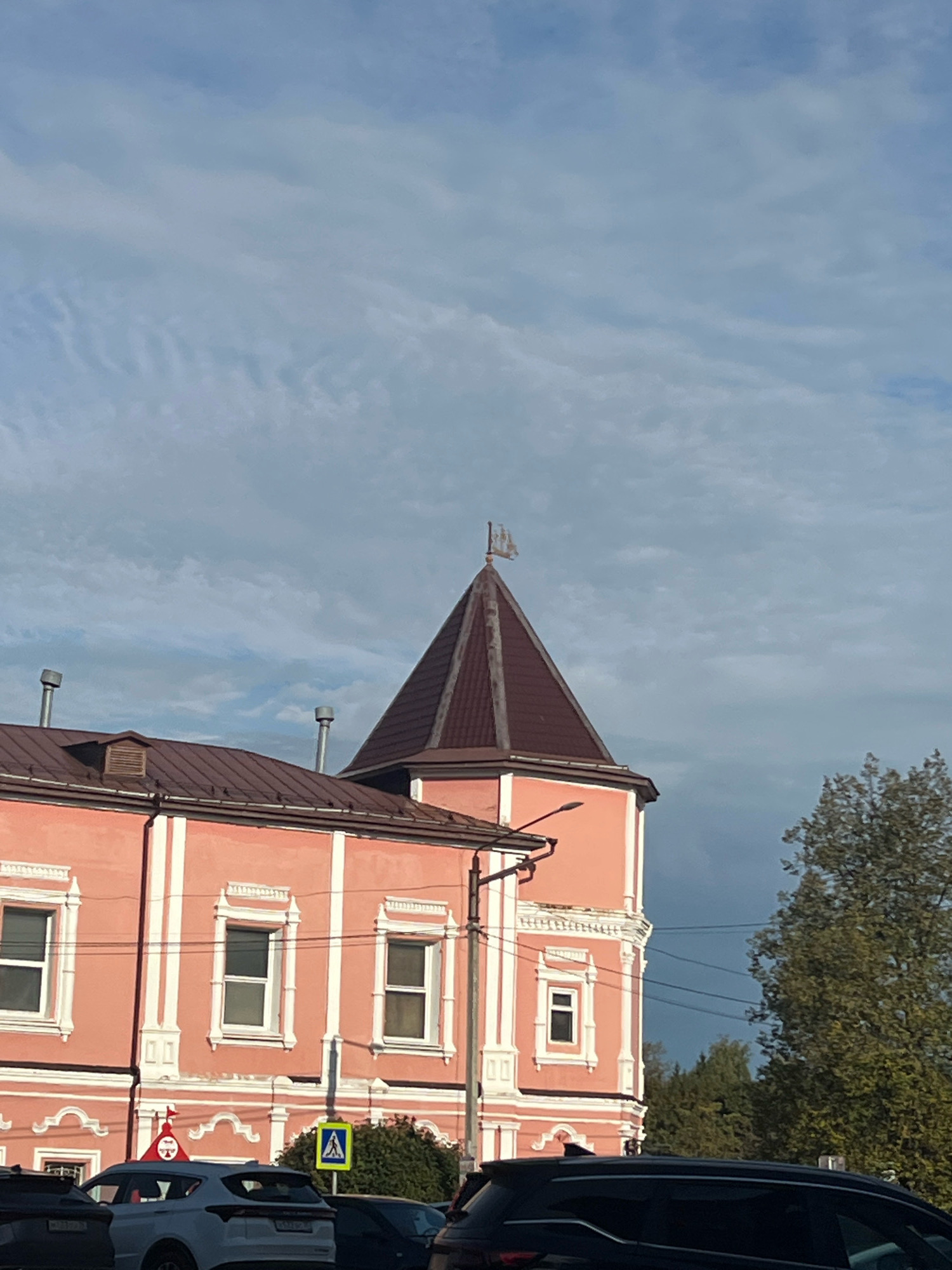 A close-up, low-angle shot captures a pink, three-story building with white trim and decorative window frames, topped by a brown pyramidal roof and a small tower featuring a weather vane shaped like a ship. Several car windows and roofs are visible in the foreground. The sky above is a mix of blue with scattered, wispy clouds, and a large green tree is visible to the right. The image appears to have been taken on a cloudy day.