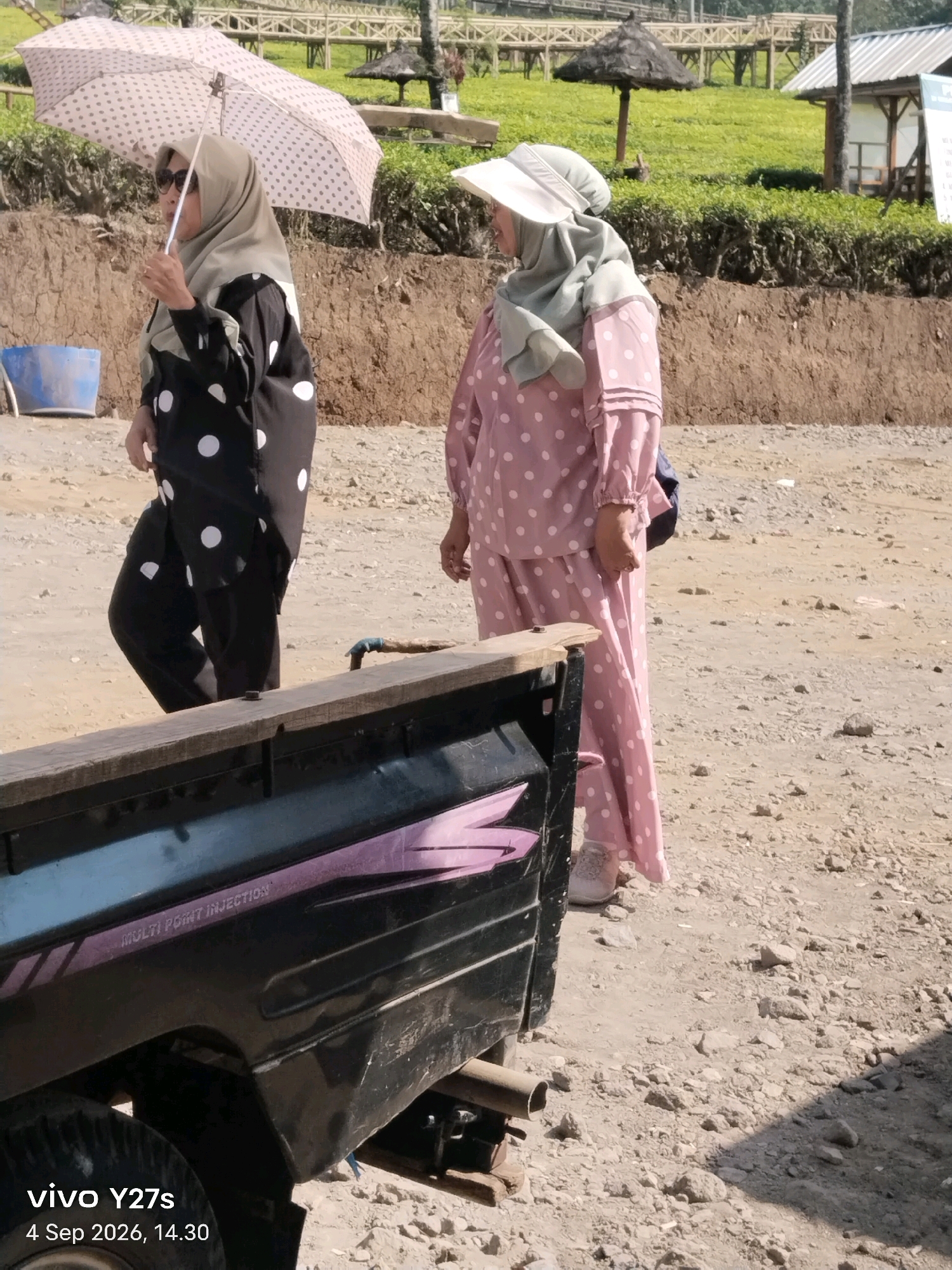 Two women are standing outdoors in a rural area, possibly Sigedang, Indonesia. The woman on the left is holding a white umbrella with a pink polka dot pattern and wearing sunglasses, a light-colored hijab, and a black garment with large white polka dots. The woman on the right is wearing a light-colored hat, a light green hijab, and a pink polka dot outfit. They are standing on a dirt and gravel surface. In the foreground, the back of a black vehicle is visible, with the text "MULTI POINT INJECTION" printed on its side. In the background, there is a grassy hillside with some vegetation and structures, including a wooden walkway and thatched roofs, suggesting a park or tourist area. The date and time stamp "vivo Y27s 4 Sep 2026, 14.30" is superimposed on the lower left corner of the image.