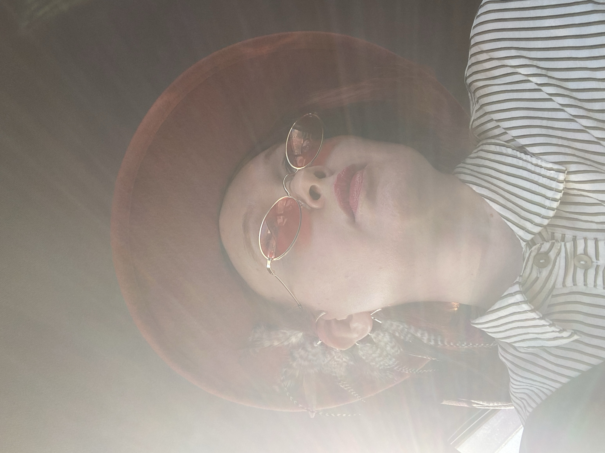 A portrait photograph, rotated counter-clockwise, features a person, likely a woman, seen from a slightly low angle. She is wearing a wide-brimmed, reddish-brown hat and small, round, gold-rimmed sunglasses with reddish lenses. Her fair skin is visible, along with red lipstick on her lips. She is dressed in a white shirt with thin horizontal brown stripes, featuring visible buttons. Decorative feathers, possibly attached to the hat, are visible near her right ear.
A strong, bright lens flare emanates from the lower-left, casting a significant haze and glow across much of the image, suggesting bright, possibly direct sunlight. The background is dark and indistinct, offering no specific environmental cues. The person's expression is neutral, with her eyes obscured by the sunglasses. No activity is depicted beyond posing. No visible text is present. The scene is set in Yekaterinburg, Russia.