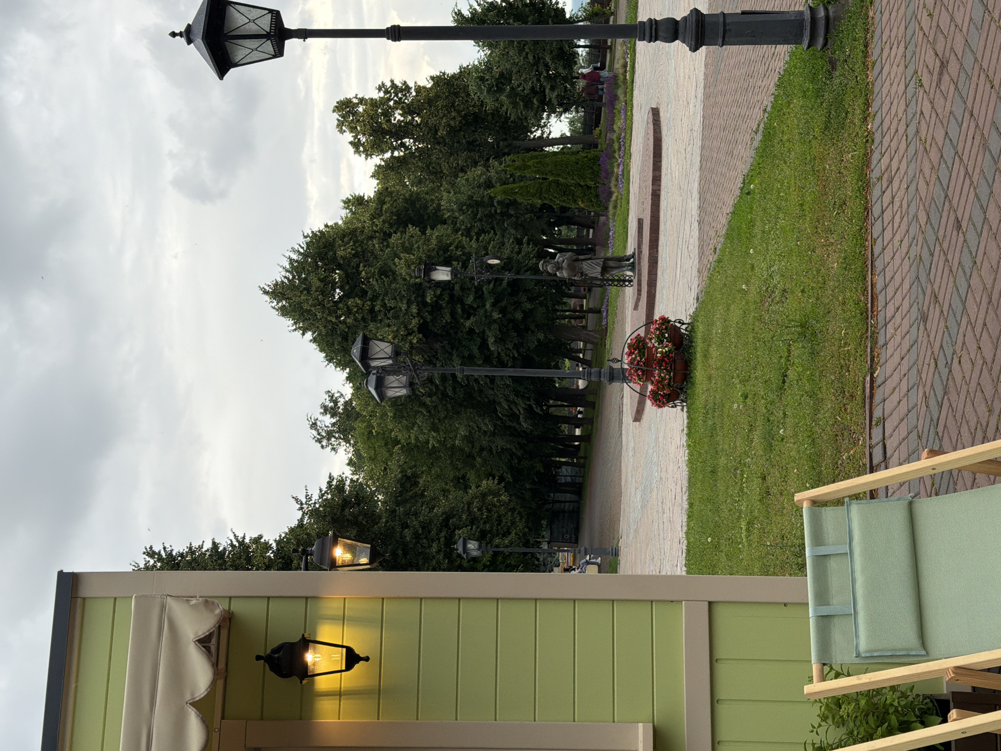 A vertical, slightly tilted outdoor medium shot captures a park scene on an overcast day. The foreground features the top of a light green building with a scalloped cream awning and two decorative black lanterns, one of which is lit. To the right, a portion of a light green canvas lounge chair with a wooden frame is visible.
The mid-ground shows a paved walkway made of light and dark grey bricks, bordered by a strip of green grass. A black lamppost stands on the left side of the walkway. Further back, a row of dark green trees lines the path, with hints of lavender flowers peeking through. A small statue of two people, possibly a couple, stands near the lamppost and in front of the trees. A few people are seated on a bench in the distance. The background is dominated by a cloudy, grey sky.
The scene suggests a relaxed, possibly late afternoon or early evening atmosphere in a public park in Ulyanovsk, Russia. The cloudy weather contributes to a subdued mood. The lighting on the lanterns indicates it is getting dark or is already dim. There is no visible text in the image.