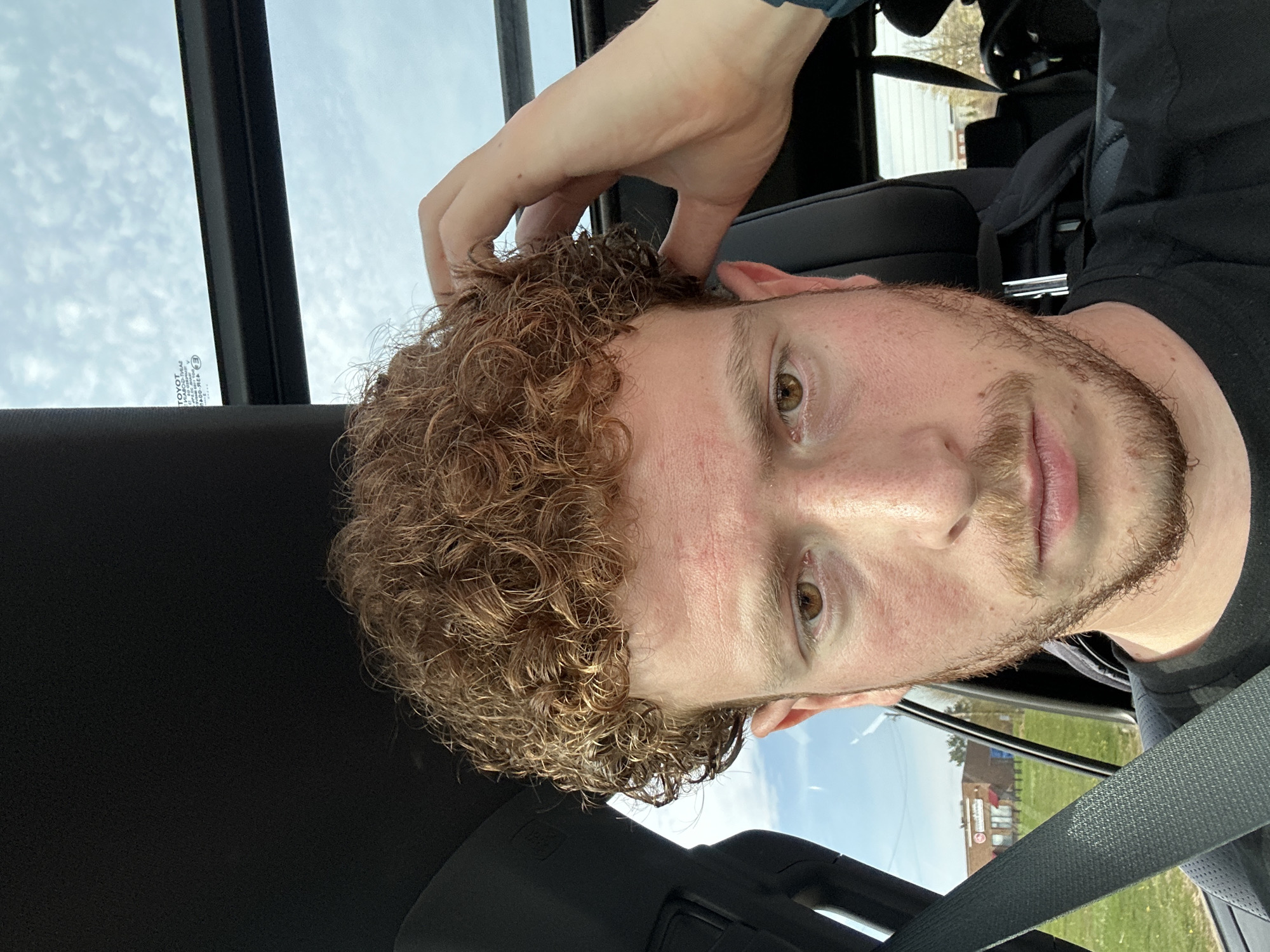 A person with curly, light brown hair and a beard is shown in the driver's seat of a car. The person is looking directly at the camera with their right hand touching their hair. The car's interior is visible, including the dashboard and window. The background shows a building in the distance, possibly located in Beachwood, United States of America. The image is taken from a low angle, looking up at the person. The person is wearing a seatbelt and a dark-colored shirt. There's a sky visible through the car's window.
