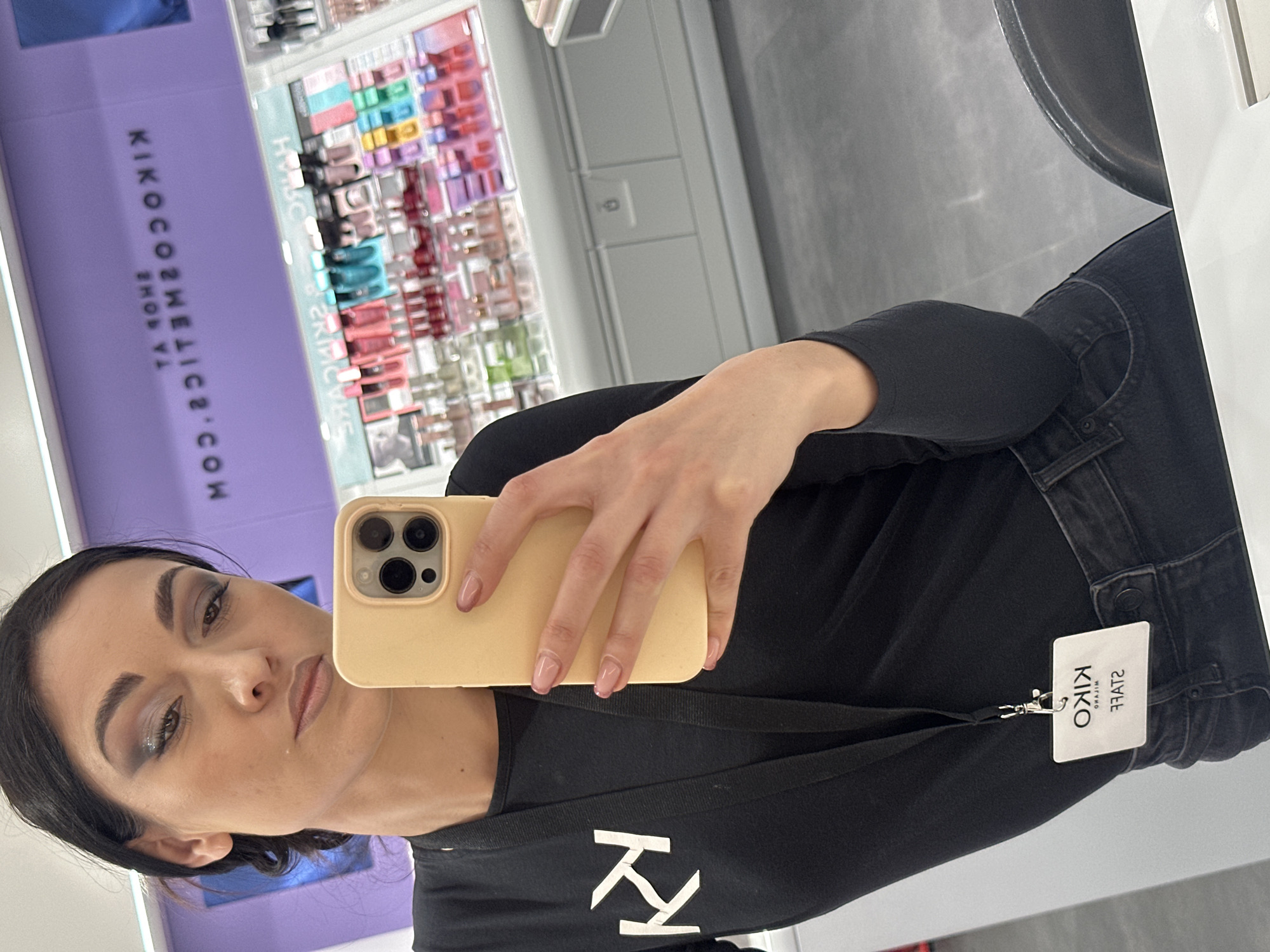 A woman is taking a selfie in what appears to be a cosmetics store. She is holding a yellow iPhone, obscuring part of her face. The woman has dark hair pulled back, with distinct eye makeup. She is wearing a black shirt with a white logo that appears to be 'KK', and a 'KIKO STAFF' tag is visible near her denim jeans. In the background, there is a display of various cosmetics, and the store's signage reads "KIKOCOSMETICS.COM". The exact location within the city of Unknown, Unknown cannot be determined from the image.