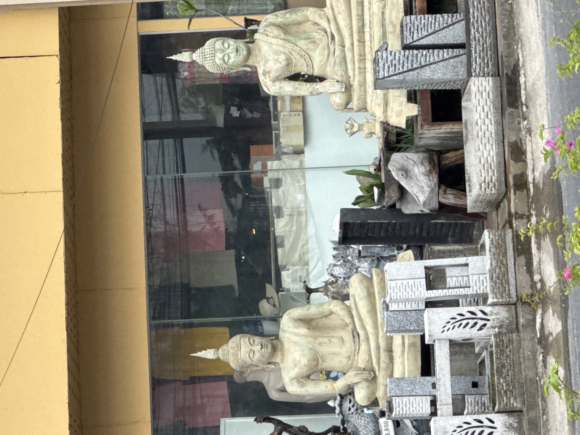 In Kathu, Thailand, a storefront displays religious statues and stone carvings. Two large, light-colored Buddha statues in meditation are prominently featured. The statue on the left is positioned behind an array of smaller stone decorative items, including planters with leaf motifs and multi-tiered stone structures. The statue on the right, also seated in meditation, is slightly more weathered and appears to be in front of a display of what might be Buddhist flags or banners within the shop window. A headless mannequin is visible behind the left Buddha statue, along with a straw hat on a shelf. Various other stone sculptures and decorative objects are placed around the statues and in front of the storefront, suggesting a retail environment for religious or garden ornaments. The storefront has a yellow awning and a large glass window reflecting the items inside and the exterior environment. Small pink flowers are visible in the foreground, indicating an outdoor setting.