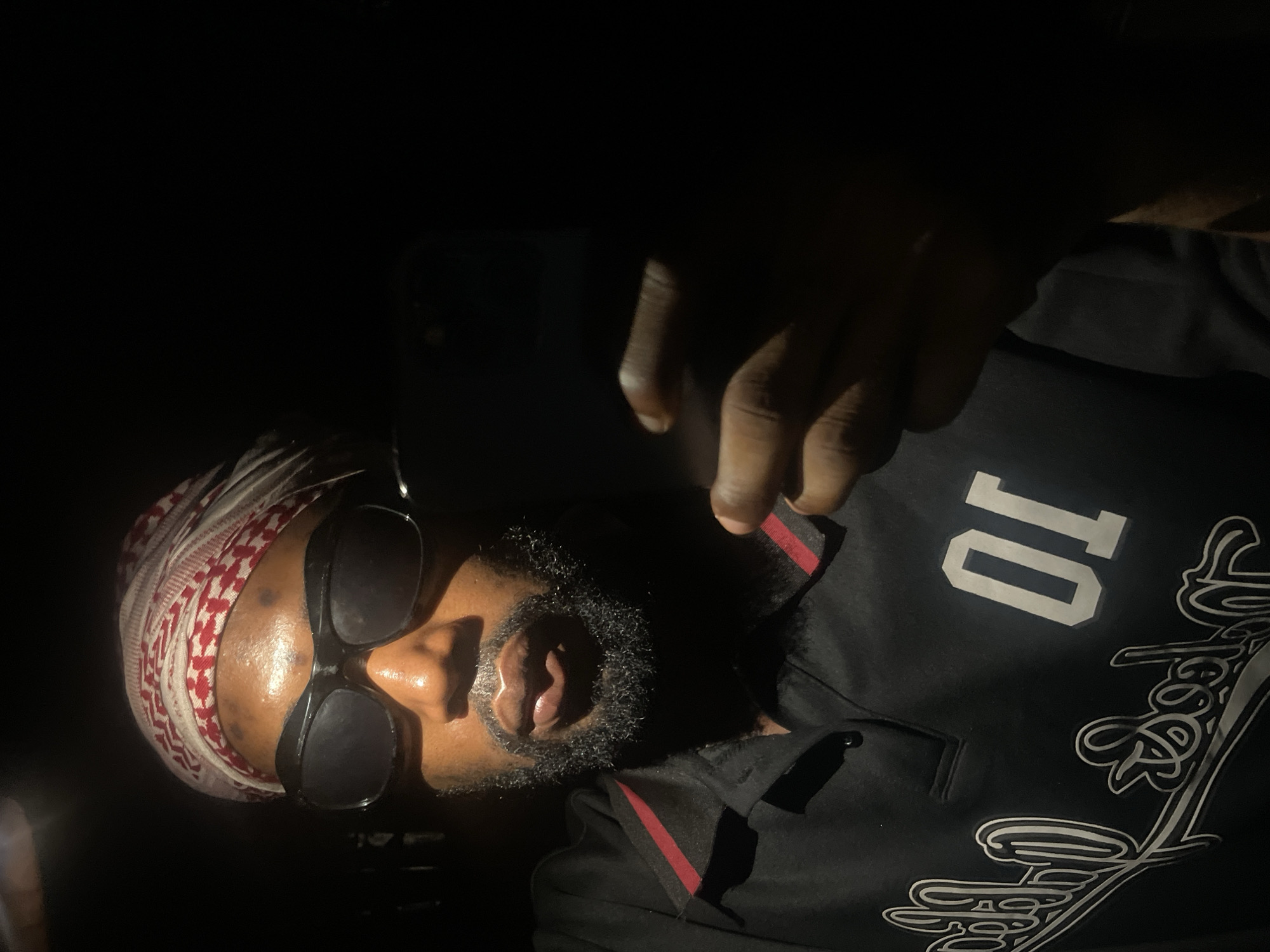 A man is seen wearing a red and white patterned head covering and dark sunglasses. He has a full beard. He is wearing a black shirt with a red stripe on the collar and a white "10" on the shoulder. A hand is visible above his head. The photograph appears to be taken in dim lighting. There is no information to suggest this photograph was taken in Lokoja, Nigeria.