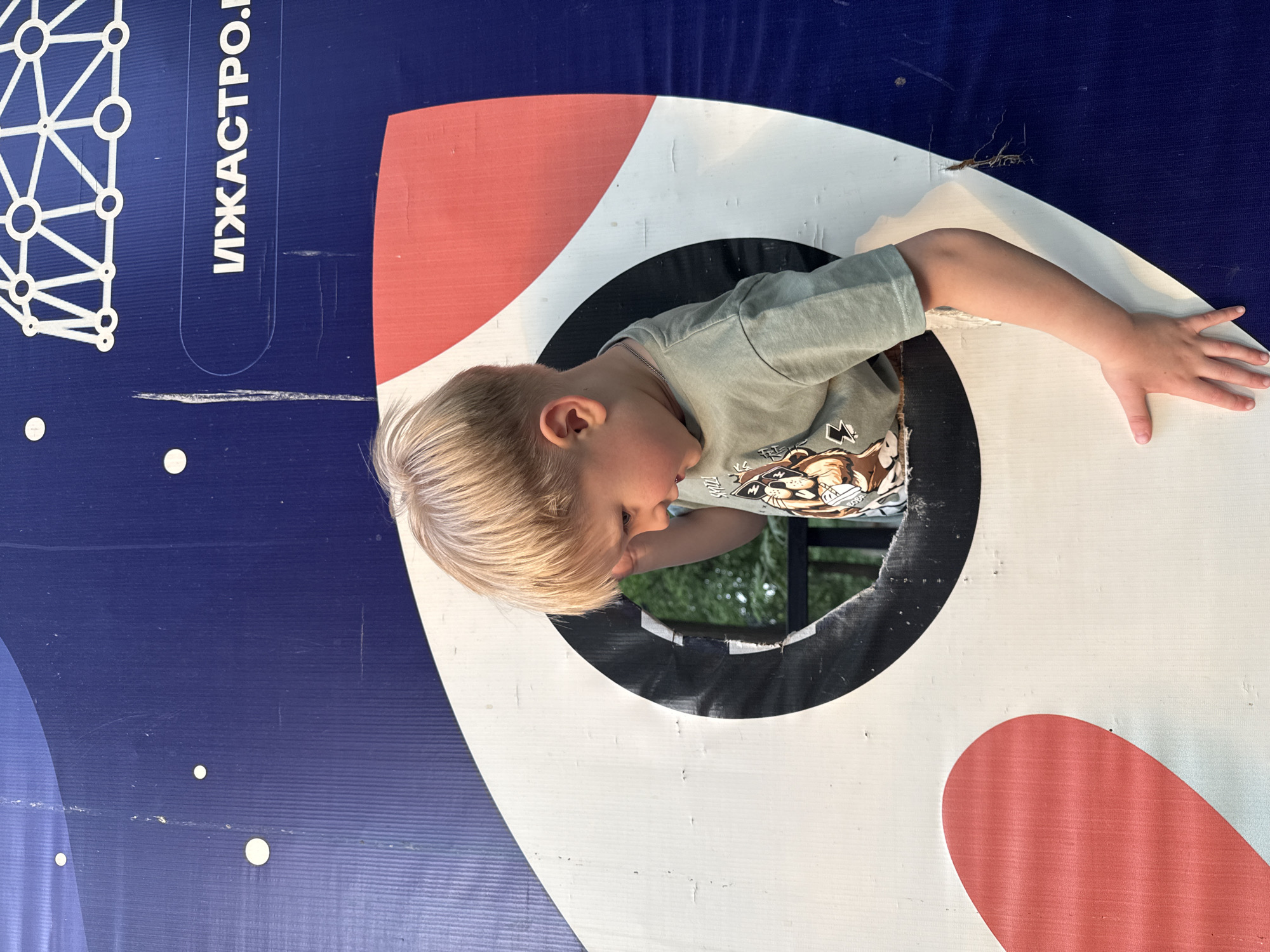A young boy with blonde hair is looking through a hole in a rocket-shaped cutout. The rocket is painted white with red fins and is set against a blue background with white dots and a white geometric design. Inscribed on the blue area is the text "ИЖАСТРО". The boy is wearing a green t-shirt with a graphic on it. Behind the cutout, greenery is visible. This scene is likely in Izhevsk, Russia.