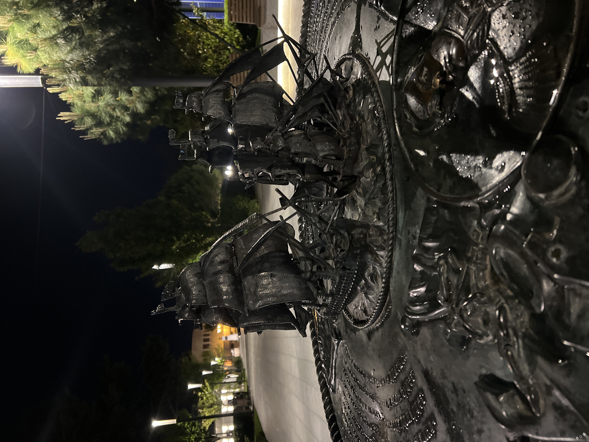 The image depicts a monument, likely made of bronze or another dark metal, in what appears to be an outdoor public space in Sevastopol, Ukraine. The monument features detailed ship models, potentially representing historical naval vessels. The ships are mounted on a circular base, possibly a depiction of a compass rose or another nautical symbol. The monument is illuminated by artificial light sources, including streetlights, which contribute to the nighttime ambiance. In the background, there are trees and buildings, typical of an urban park setting. The image is taken at close range, focusing on the details of the monument's craftsmanship.
