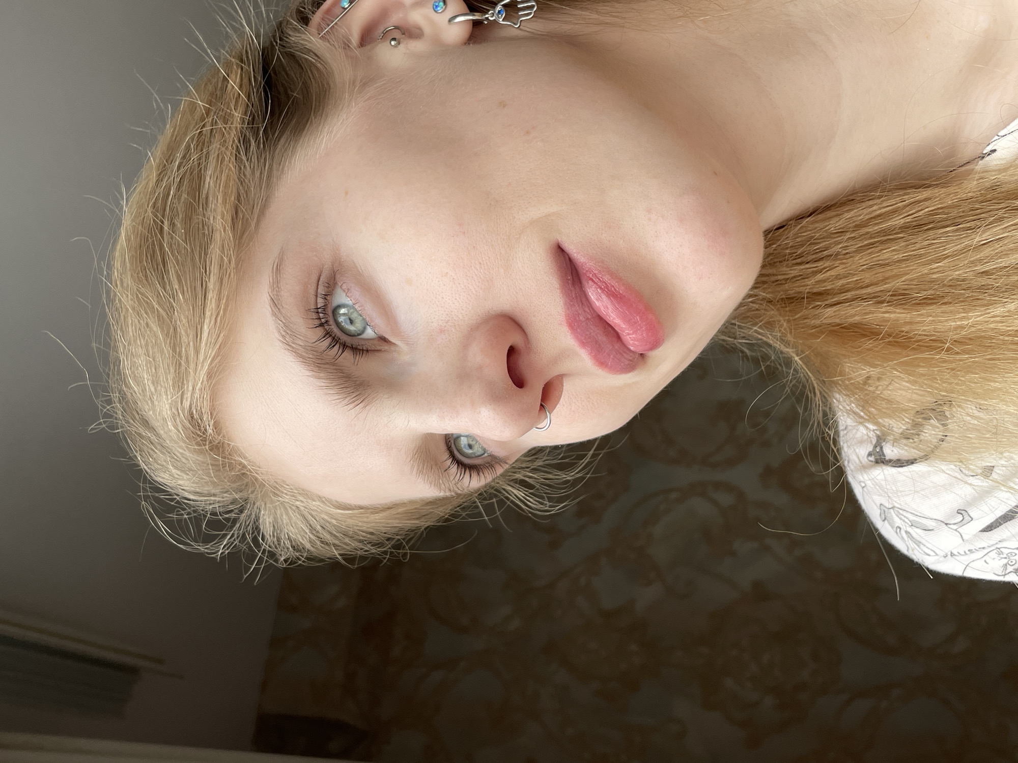 A close-up, tilted portrait shows a fair-skinned person with light blonde hair and light blue eyes. They have multiple piercings in their left ear, including a helix piercing with a turquoise stud, a conch piercing, and an industrial piercing. A silver ring is in their left nostril. Their lips are slightly parted, revealing the tongue. The background is a patterned wall, with a partial view of a window or blinds in the lower left. The person is wearing a white garment, possibly a t-shirt, with black line art visible. No information regarding the location in the city of Izhevsk, Russia is available from the image.