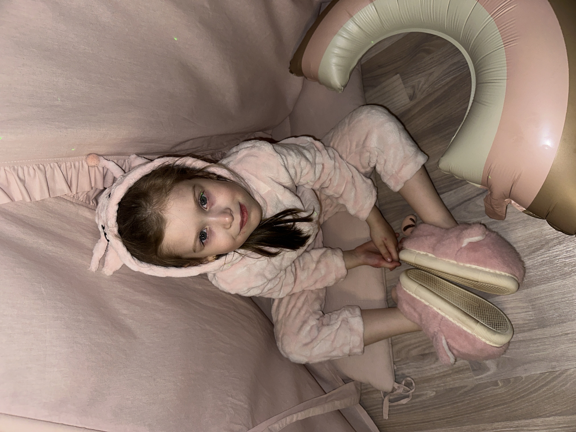 A young girl is depicted lying down in an indoor setting, likely in a cozy home environment in Chaikovsky, Russia. She is the primary subject, shown looking directly at the camera with a gentle smile and bright blue-green eyes. She wears a soft, fluffy pink animal-themed onesie with a hood that features small ears, and matching pink, plush slippers. Her dark hair is visible peeking from under the hood.
The girl is positioned on a surface covered with light pink fabric, which has a ruffled edge, suggesting it could be a bed, a play tent lining, or a large cushion. To her right, a portion of a large inflatable ring is visible, displaying stripes of beige, light pink, and a darker, possibly metallic brown or gold color, resting on a light-colored wooden floor. The overall scene conveys a sense of comfort and relaxation, possibly during leisure time or before bed. The lighting is soft, consistent with an indoor evening or night setting. No visible text is present in the image.