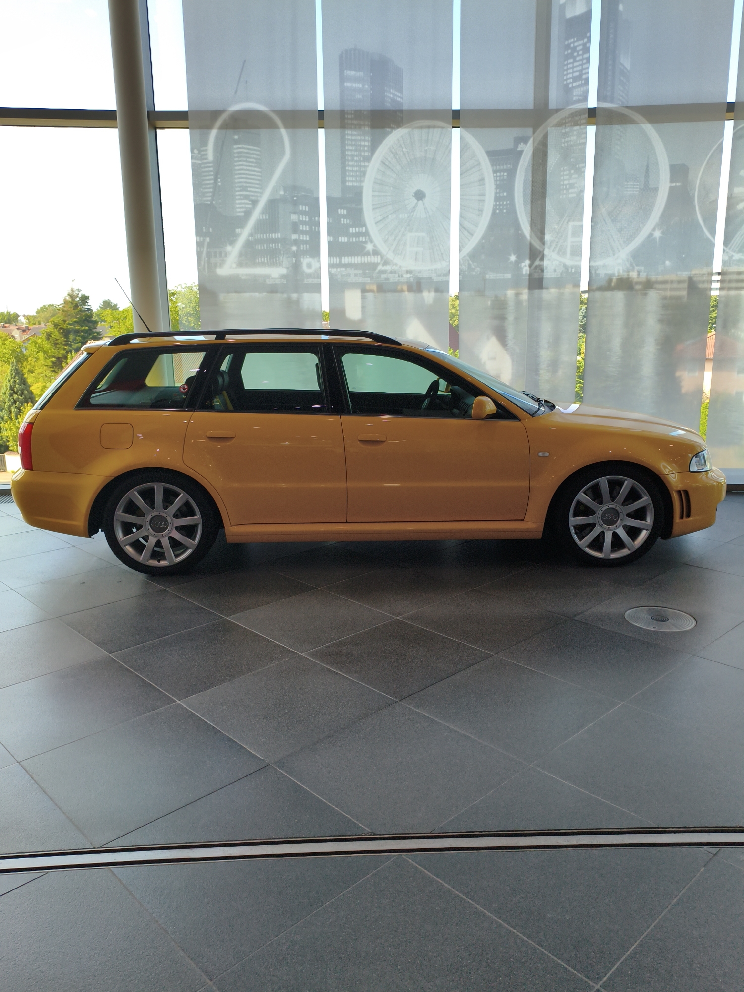 An orange Audi RS4 Avant is parked indoors on a tiled floor, likely in a showroom or museum setting. The car is viewed from the side, showcasing its wagon body style. Behind the car, sheer white curtains are drawn, featuring a faint, large-scale graphic of ferris wheels and buildings, suggesting an urban or event-related backdrop. The floor tiles are large, square, and dark grey, with faint grout lines. Sunlight streams in from the left, casting some light and shadows on the car and the floor. The environment appears to be a contemporary, well-lit space, possibly a part of the Audi Forum Ingolstadt, given the context of the Audi brand and the car model. There are no people visible in the image.