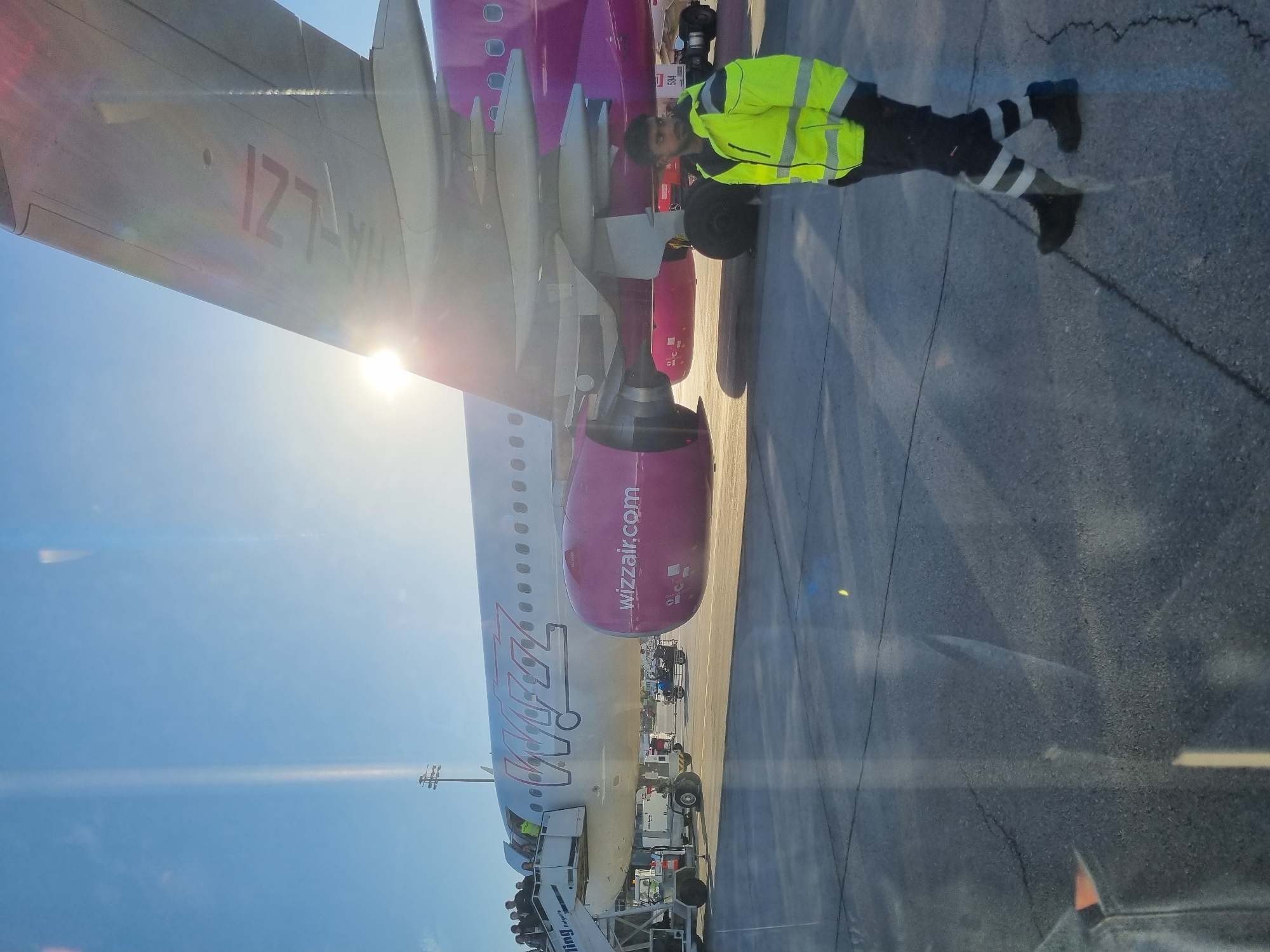The image shows a Wizz Air plane parked at a gate. An airport worker in a high-visibility vest and black pants with reflective stripes is standing near the plane's wing and engine. The registration number "HA-LZI" is visible on the wing. The Wizz Air logo is visible on the engine. The location is likely Sofia Airport, Bulgaria, given Wizz Air's presence there. The sky is blue and sunny. Airport equipment is visible near the plane. The picture was taken through glass.