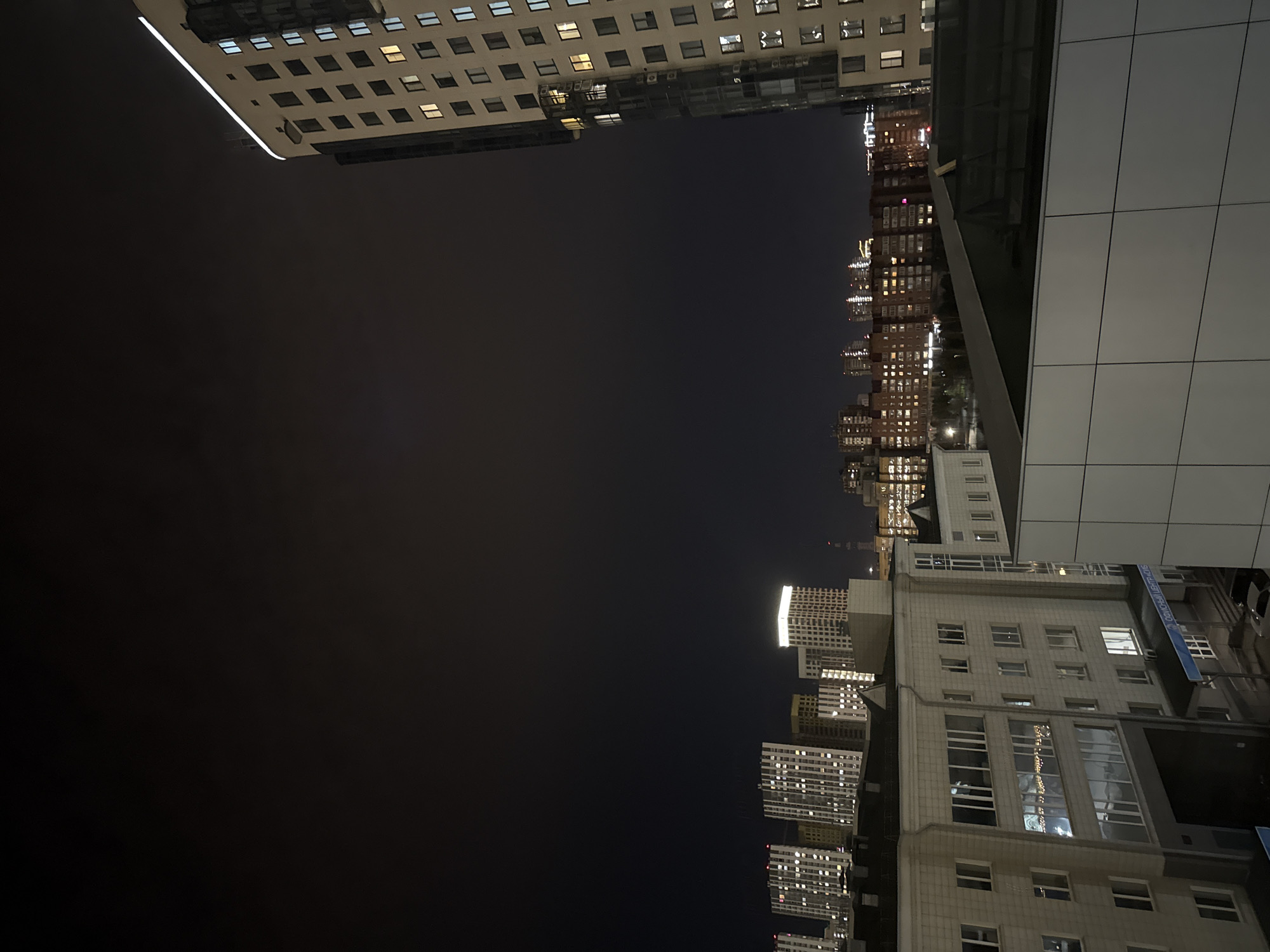 This media file captures a night-time cityscape in Krasnoyarsk, Russia, from a low-angle perspective looking upwards towards the sky and surrounding buildings. The predominant feature is a dark night sky, occupying the central and left portions of the image.
In the foreground, a prominent, modern building with a light-colored, possibly white or light gray, grid-patterned facade is visible. Its large windows reflect some light, and sections appear dark and angular, suggesting architectural details or adjacent structures.
Across the scene, numerous other high-rise buildings populate the mid-ground and background. Many of these structures have illuminated windows, indicating interior lights are on, casting a warm glow against the dark environment. The diversity of buildings ranges from more traditional designs to contemporary glass and steel structures. Decorative lights, possibly string lights, are faintly visible within the windows of one building.
The image conveys the quiet grandeur of a city at night, with artificial lights serving as the main source of illumination. No human activity is discernible.
Visible text on a blue sign attached to one of the buildings reads "ОСНОВНОЙ ЦЕНТР...", which translates to "Primary Center...". The full text is partially obscured but is consistent with a commercial or institutional building in Krasnoyarsk.